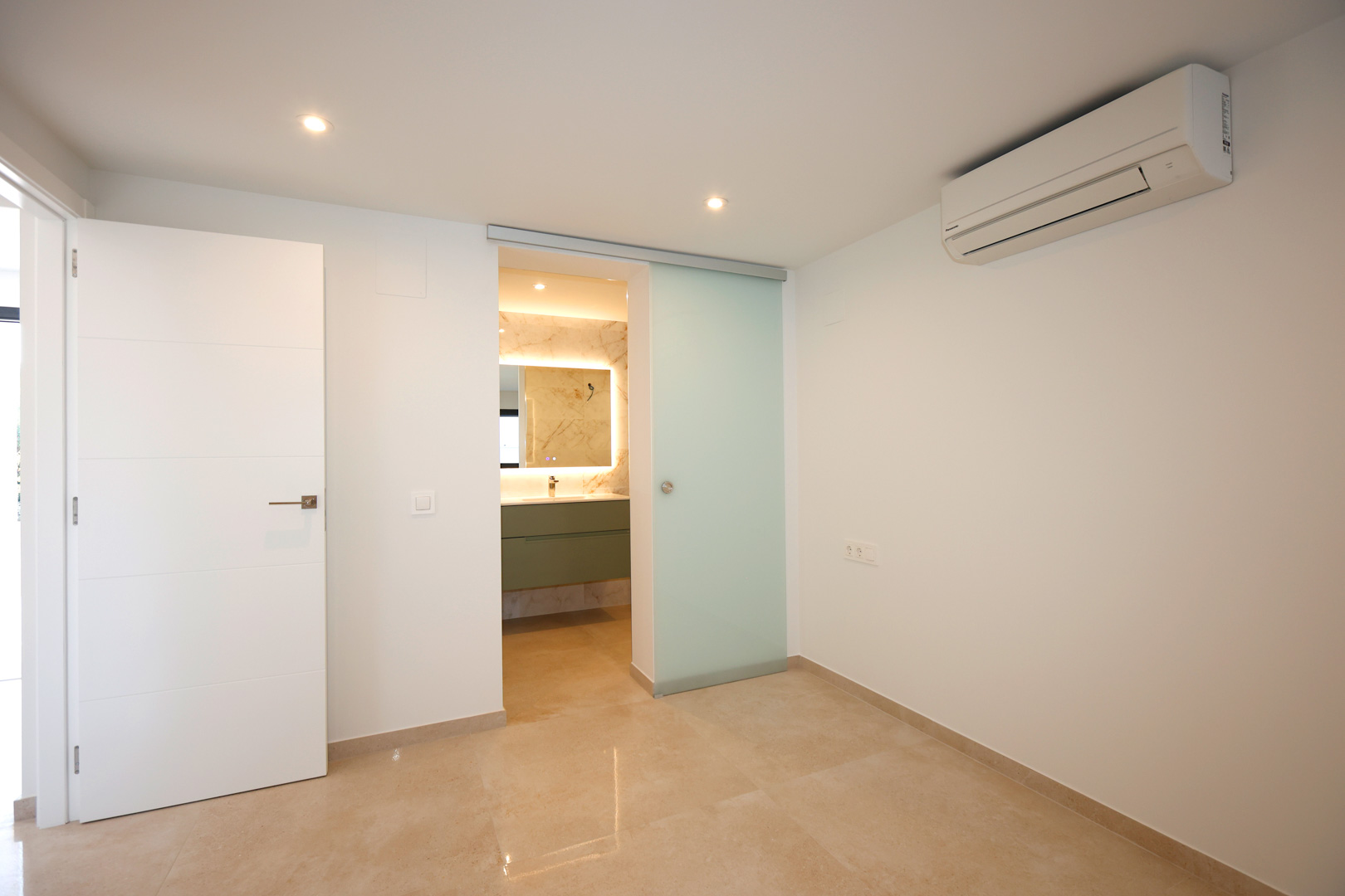 Bedroom featuring porcelain tile flooring, wall-mounted air conditioning, and a frosted glass sliding door leading to a modern en-suite bathroom.