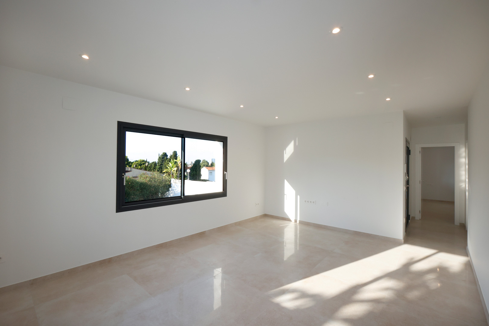 Bright living area featuring porcelain tile flooring, recessed LED lighting, and a Technal window. The hallway provides access to the entrance with an intercom system.