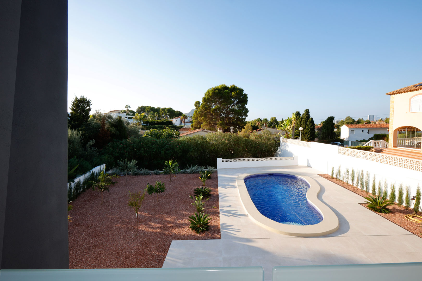Elevated view of the garden featuring a kidney-shaped pool, white porcelain tiling, and a red marble gravel area with young Mediterranean plants.