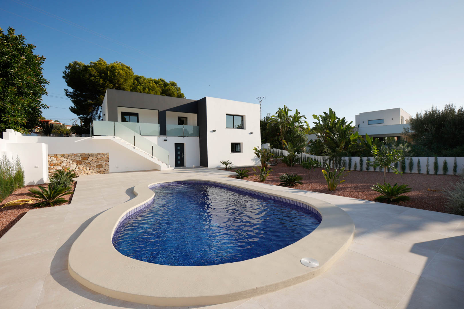 Exterior view featuring a curved swimming pool, porcelain terrace, and red marble gravel garden. The modern facade combines white and anthracite tones with glass railings.