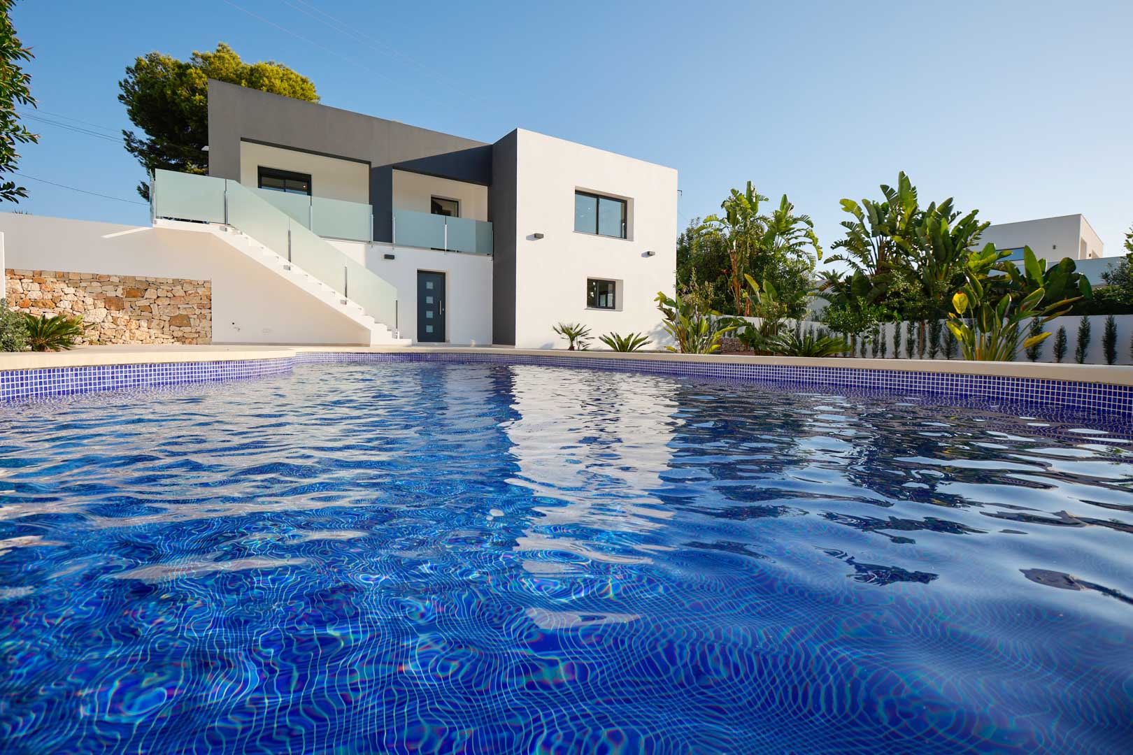 Exterior view featuring the blue tiled pool, minimalist white and grey facade, natural stone walls, and terraces with glass and steel railings.