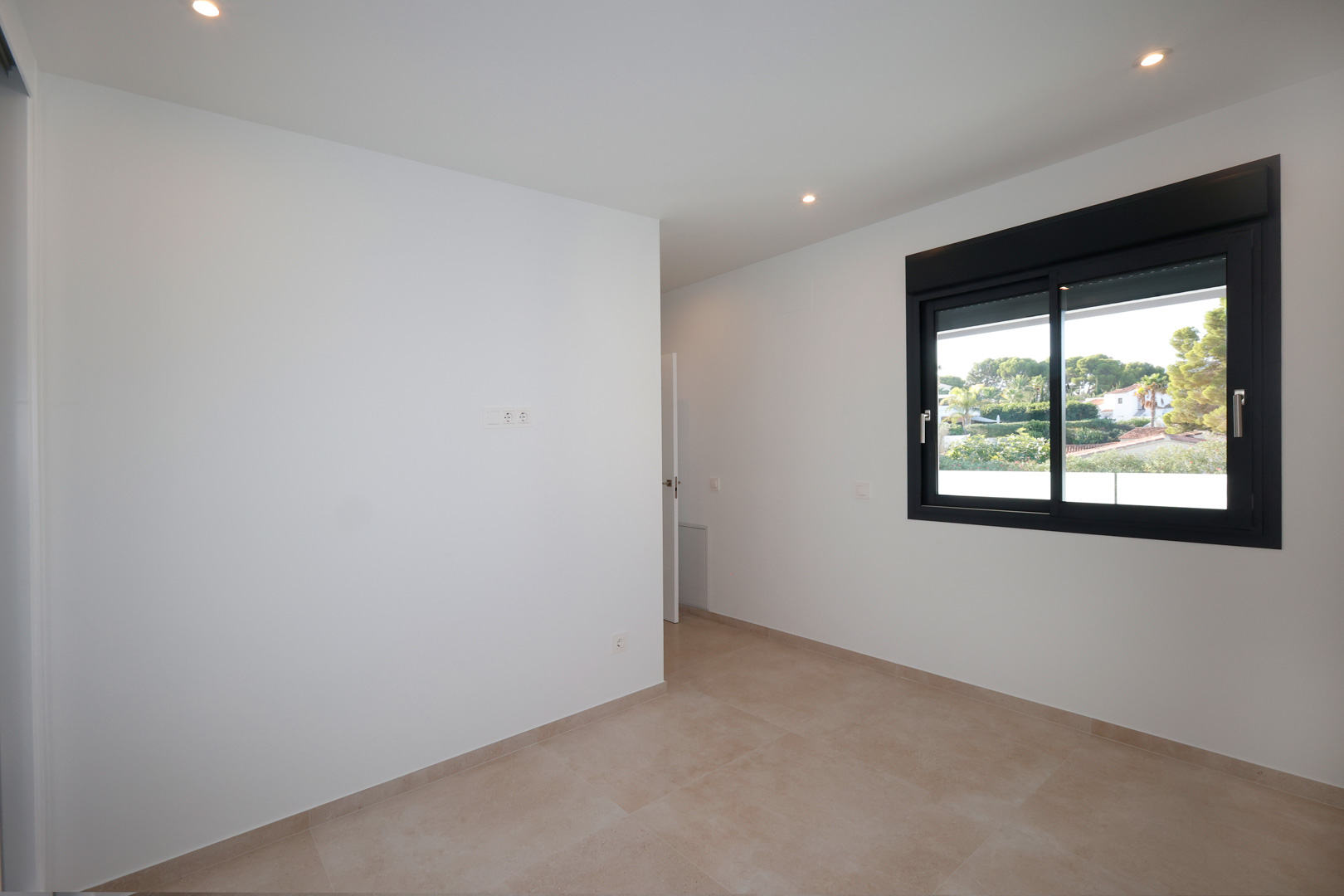 Minimalist bedroom featuring white walls, porcelain tile flooring, and a black aluminum window with electric shutters overlooking the green surroundings.