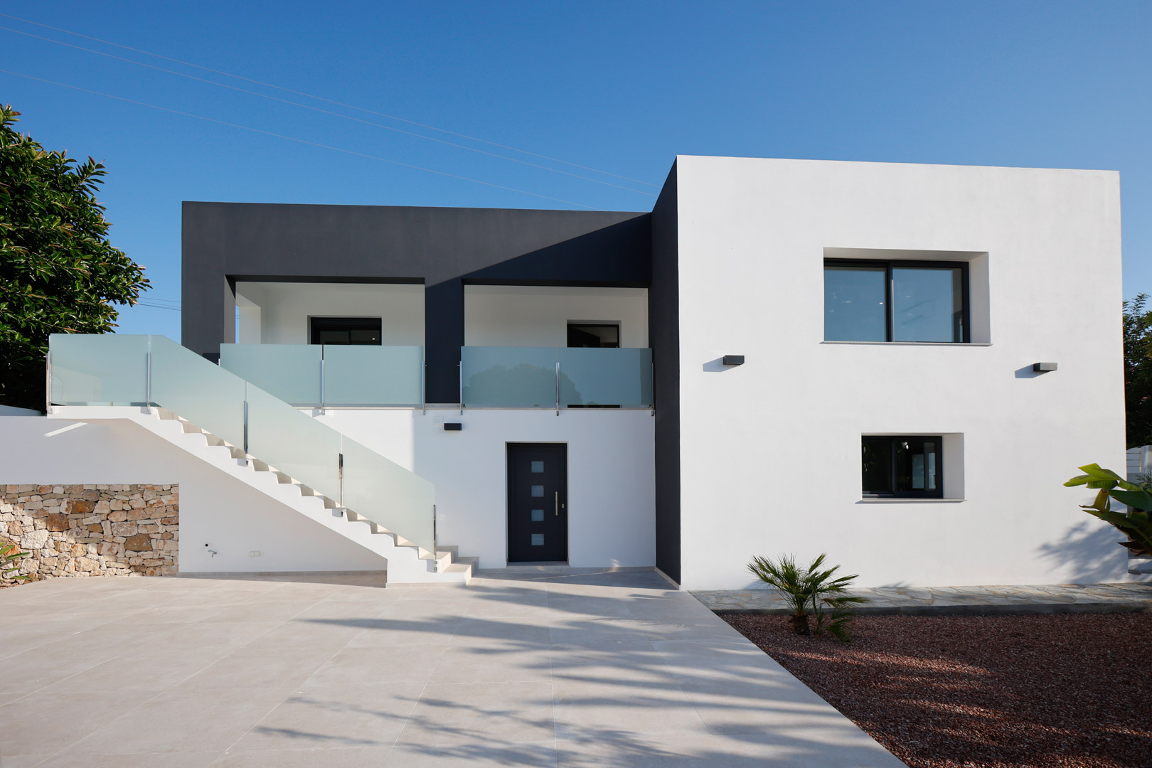 Minimalist white and grey facade featuring an external staircase with glass railings, Technal aluminum joinery, and a natural stone accent wall.