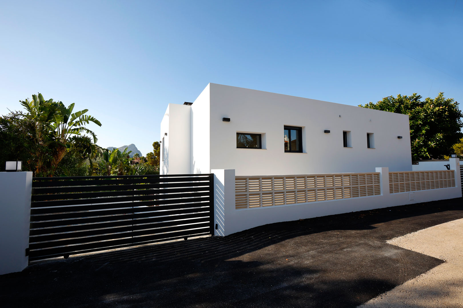 Minimalist white facade featuring a black slatted automatic gate, perimeter wall with decorative lattice, and paved access surrounded by greenery.