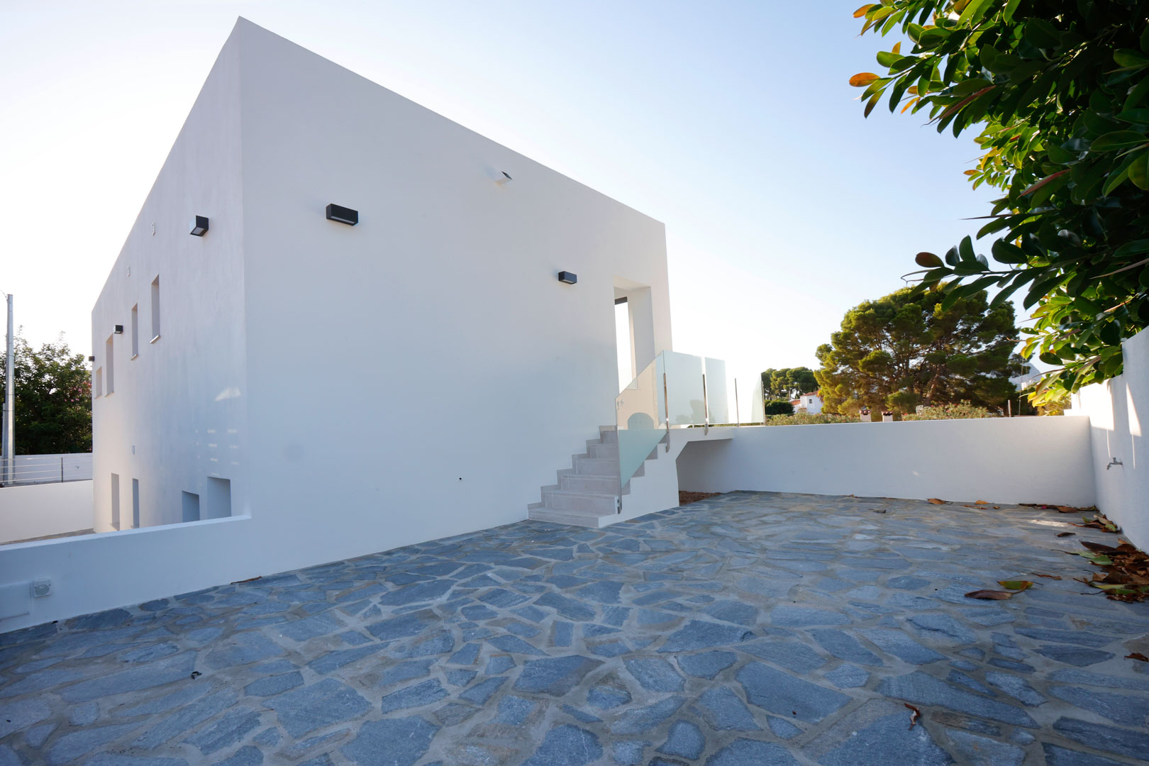 Minimalist white facade featuring a spacious irregular natural stone patio and an exterior staircase with glass and stainless steel railings.