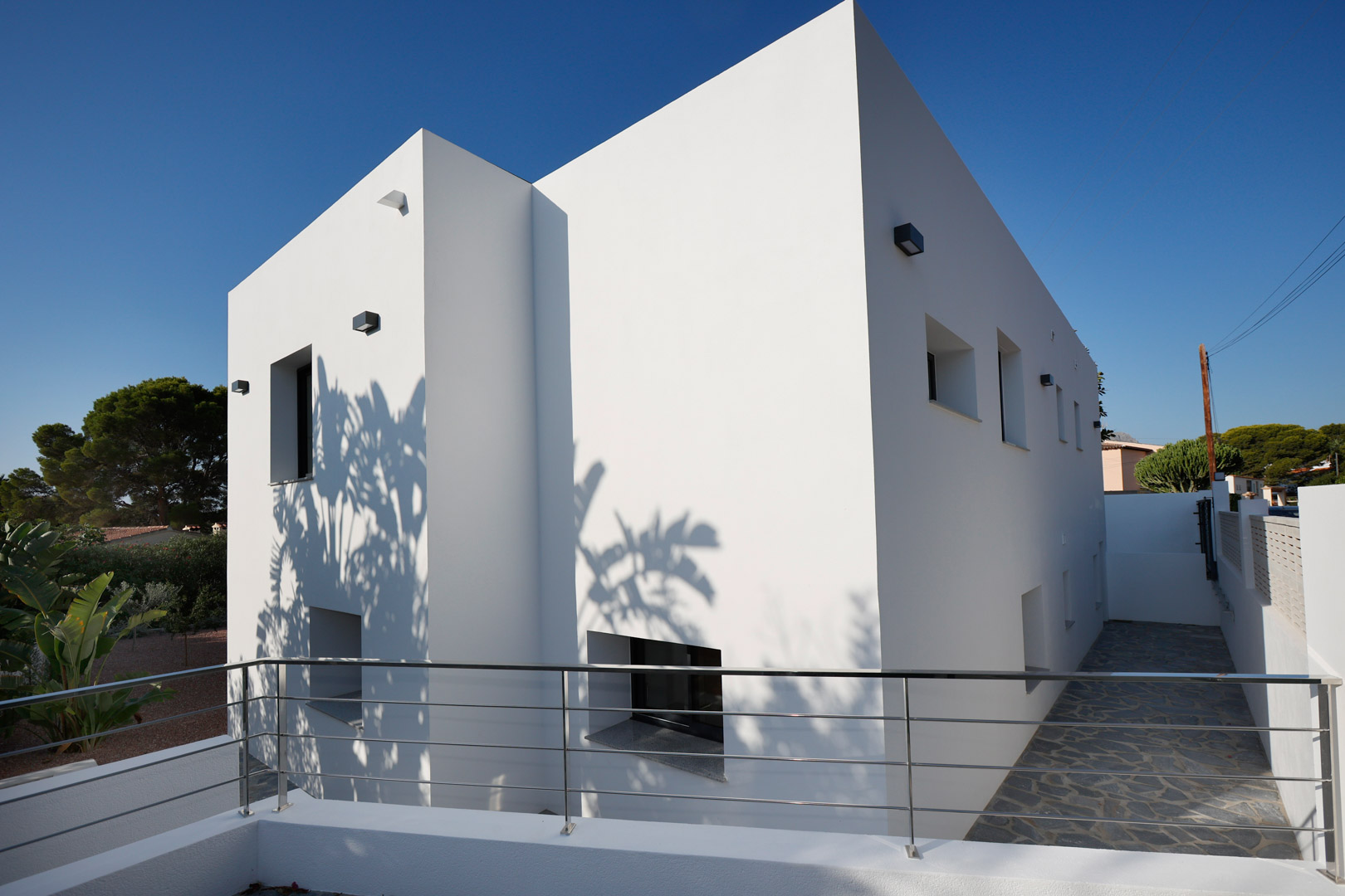 Minimalist white facade featuring clean geometric lines, stainless steel railings, and natural stone paving. Tropical shadows add a dynamic touch to the contemporary architecture.