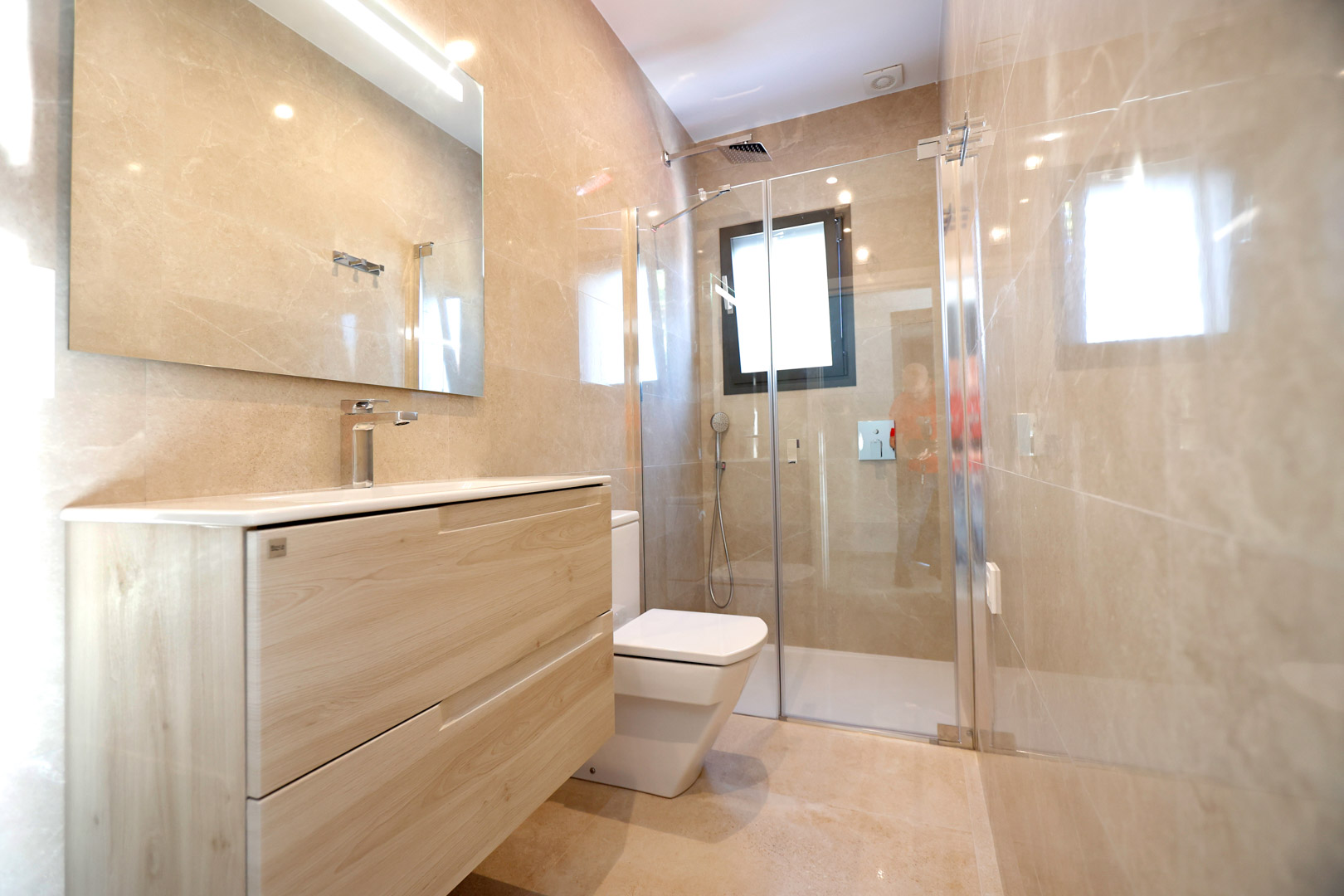 Modern bathroom with a light wood vanity, walk-in glass shower, and polished cream porcelain tiles. Features chrome fixtures and integrated mirror lighting.
