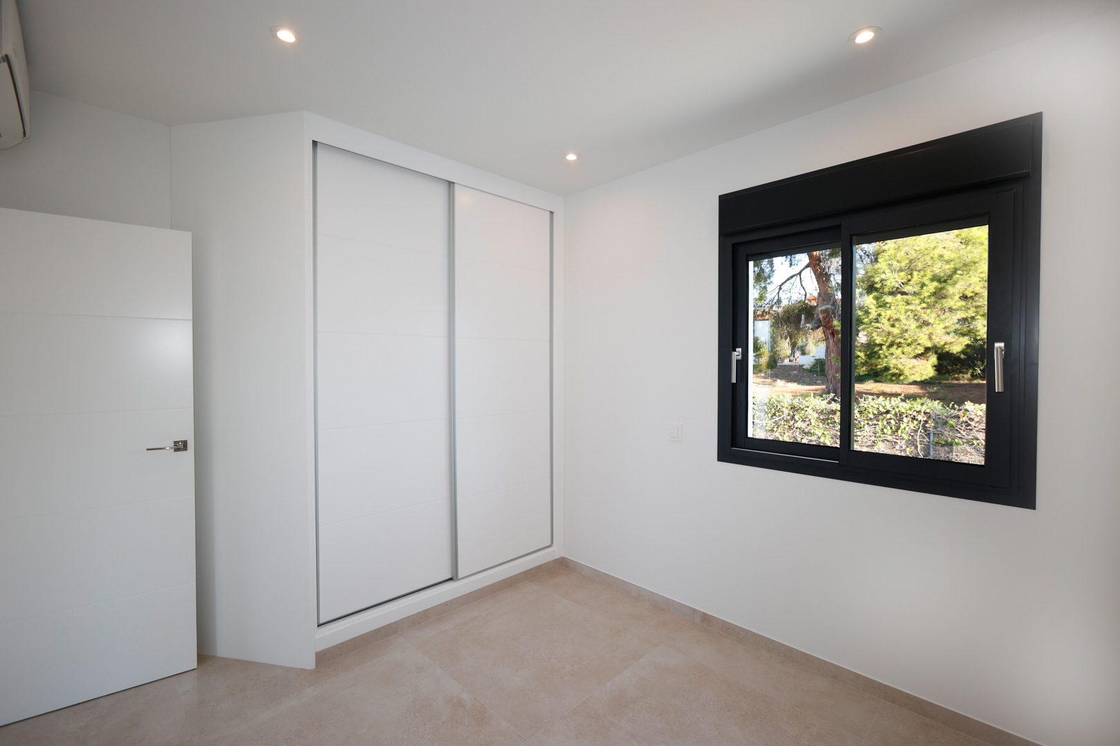 Modern bedroom featuring a white built-in sliding wardrobe, porcelain tile flooring, and a black aluminum window overlooking the garden.