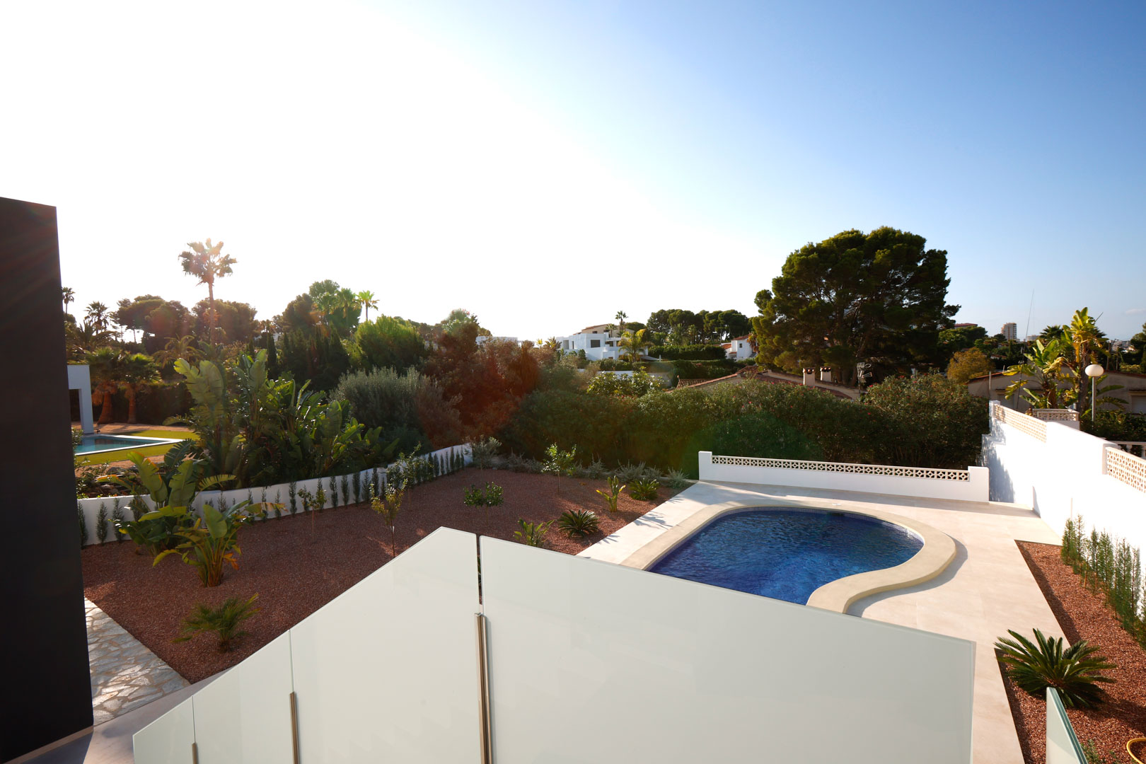 View from the upper terrace with glass railings, overlooking the curved swimming pool, red marble gravel garden, and lush Mediterranean surroundings.