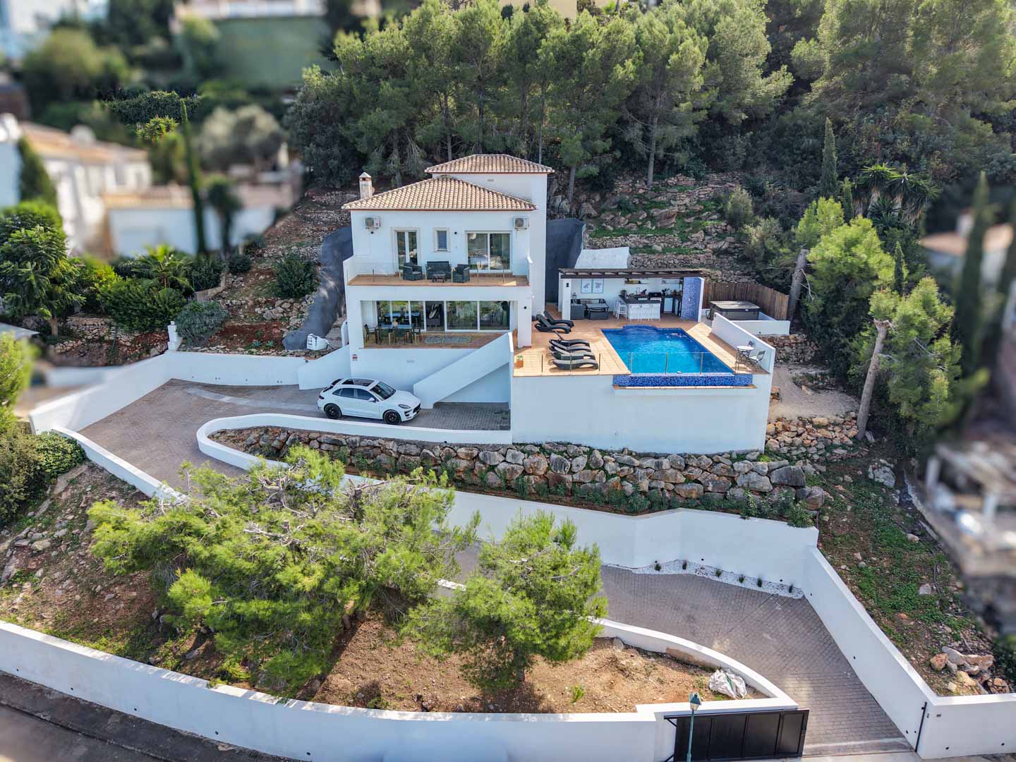 Aerial view of the Mediterranean villa featuring an infinity pool, summer kitchen, and multi-level terraces set against a pine-covered hillside in La Sella.