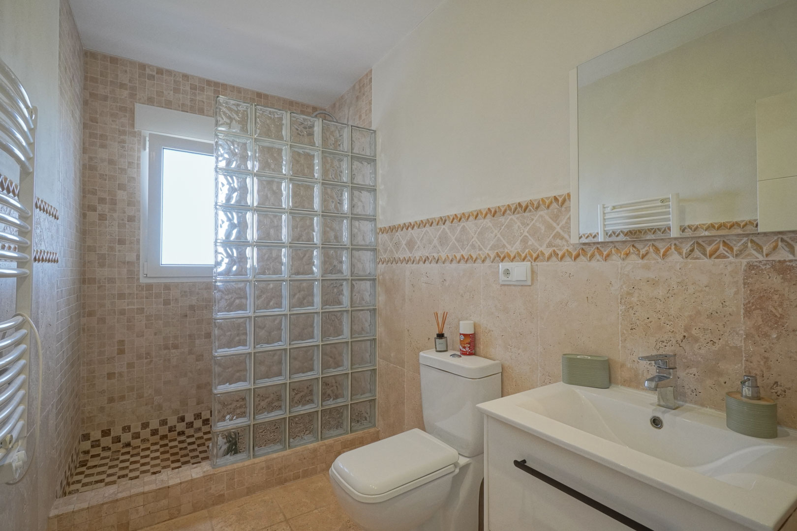 Bathroom featuring a walk-in shower with a glass block partition, earth-toned mosaic tiling, a white vanity unit, and a heated towel rail.