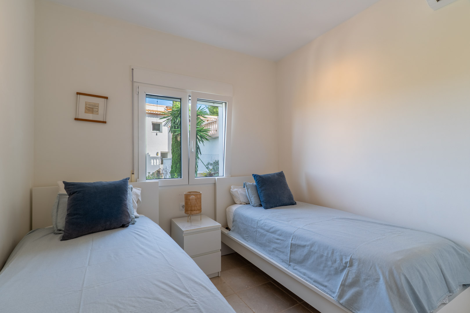Bedroom with twin beds, white frames, and a central nightstand. A PVC window provides natural light and views of the surrounding residential area.