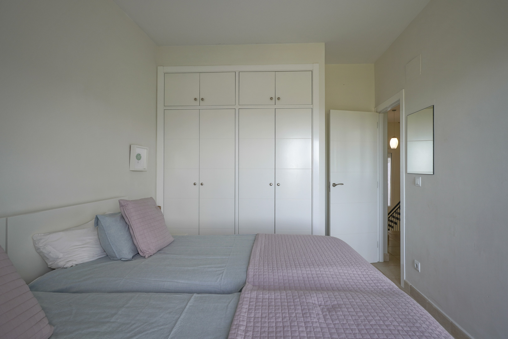 Bright bedroom featuring floor-to-ceiling white built-in wardrobes and ceramic flooring. The minimalist design emphasizes functionality and neutral tones.