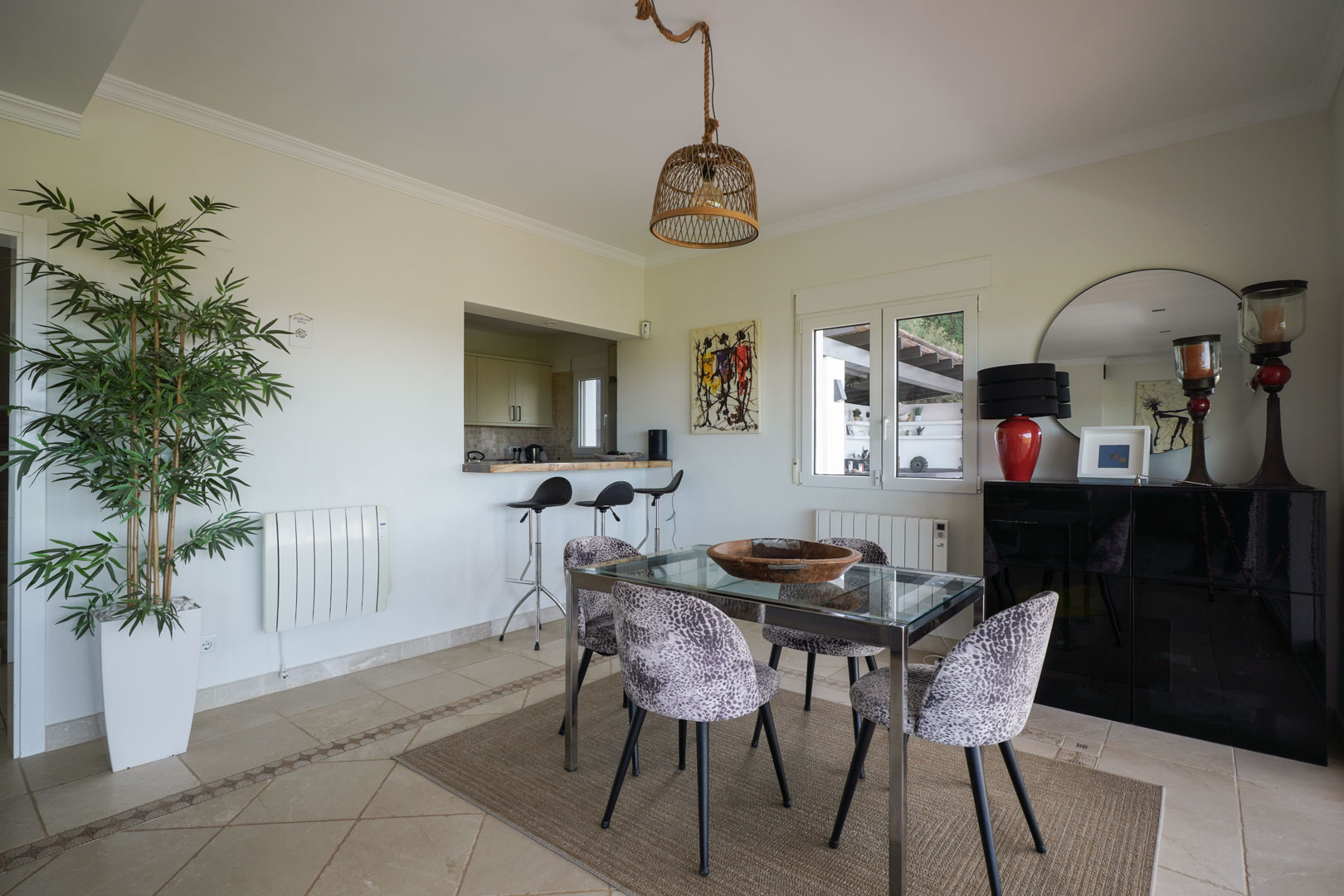 Bright dining area featuring a glass-top table, animal-print chairs, and a wicker pendant light, seamlessly connected to the kitchen via a breakfast bar.