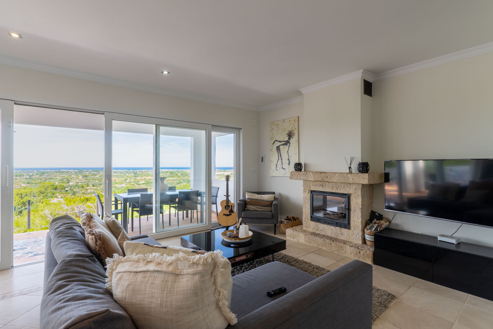 Bright living room featuring a natural stone fireplace, large sliding glass doors opening to a terrace, and panoramic views of the Mediterranean Sea and landscape.