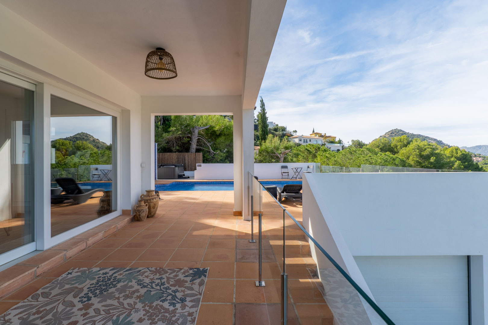 Covered terrace featuring terracotta flooring, glass balustrades, and a view of the private pool. Decorative clay pots and a rattan lamp enhance the Mediterranean style.