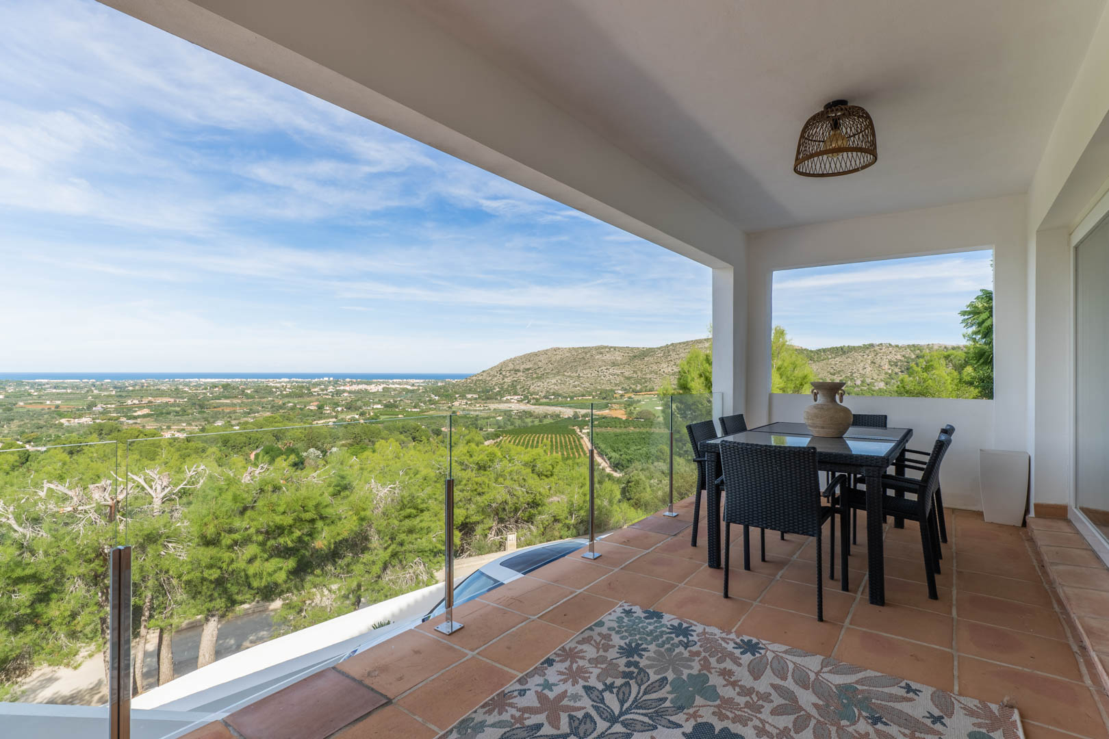 Covered terrace featuring terracotta tiles, outdoor dining set, and glass balustrades overlooking the Mediterranean Sea and lush valley landscape.