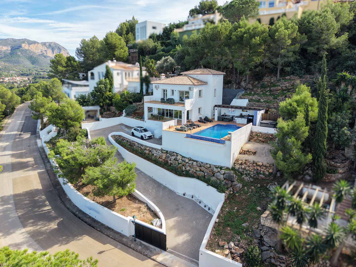 Elevated view of the Mediterranean villa featuring a private pool, multi-level terraces, and outdoor kitchen, set against a backdrop of pine trees and mountains.