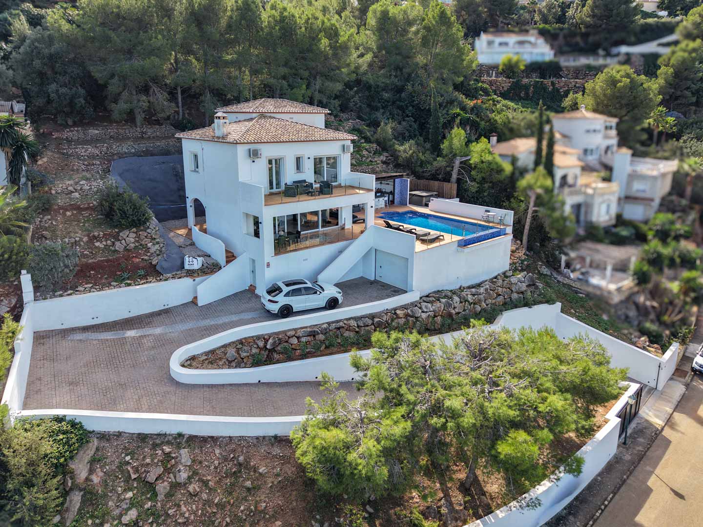 Elevated view of the villa featuring a white facade, private pool, glass-railed terraces, and a paved driveway integrated into the wooded hillside of La Sella.