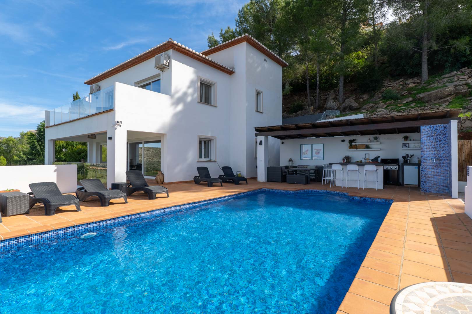 Exterior featuring a blue mosaic pool, terracotta sun deck, and a covered summer kitchen with a bar. The white villa is nestled against a natural pine-covered hillside.
