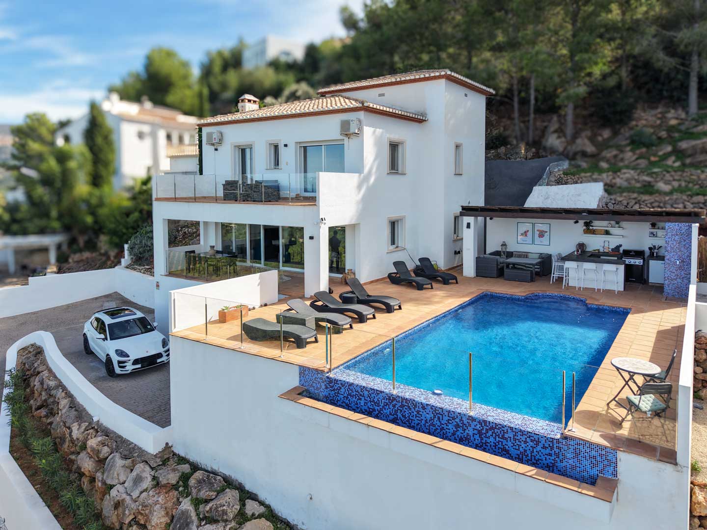 Exterior view featuring a blue mosaic pool, sun deck with loungers, outdoor kitchen under a pergola, and a white Mediterranean facade with glass-railed terraces.