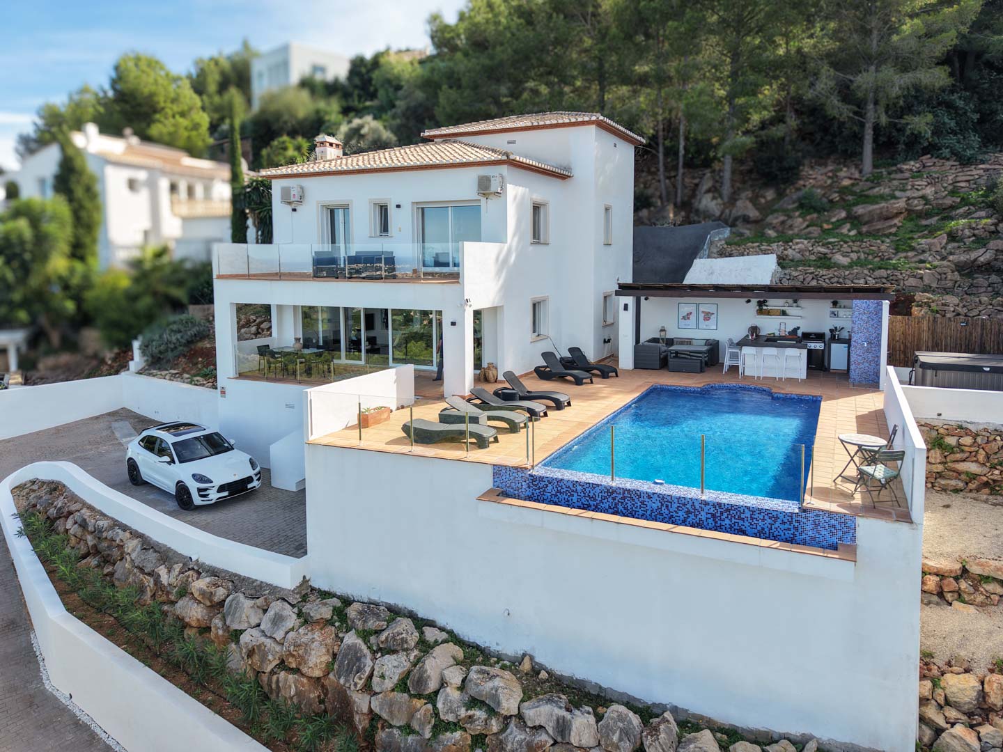 Exterior view of the villa featuring a blue mosaic pool, spacious sun deck with loungers, and a fully equipped outdoor kitchen set against a pine forest backdrop.