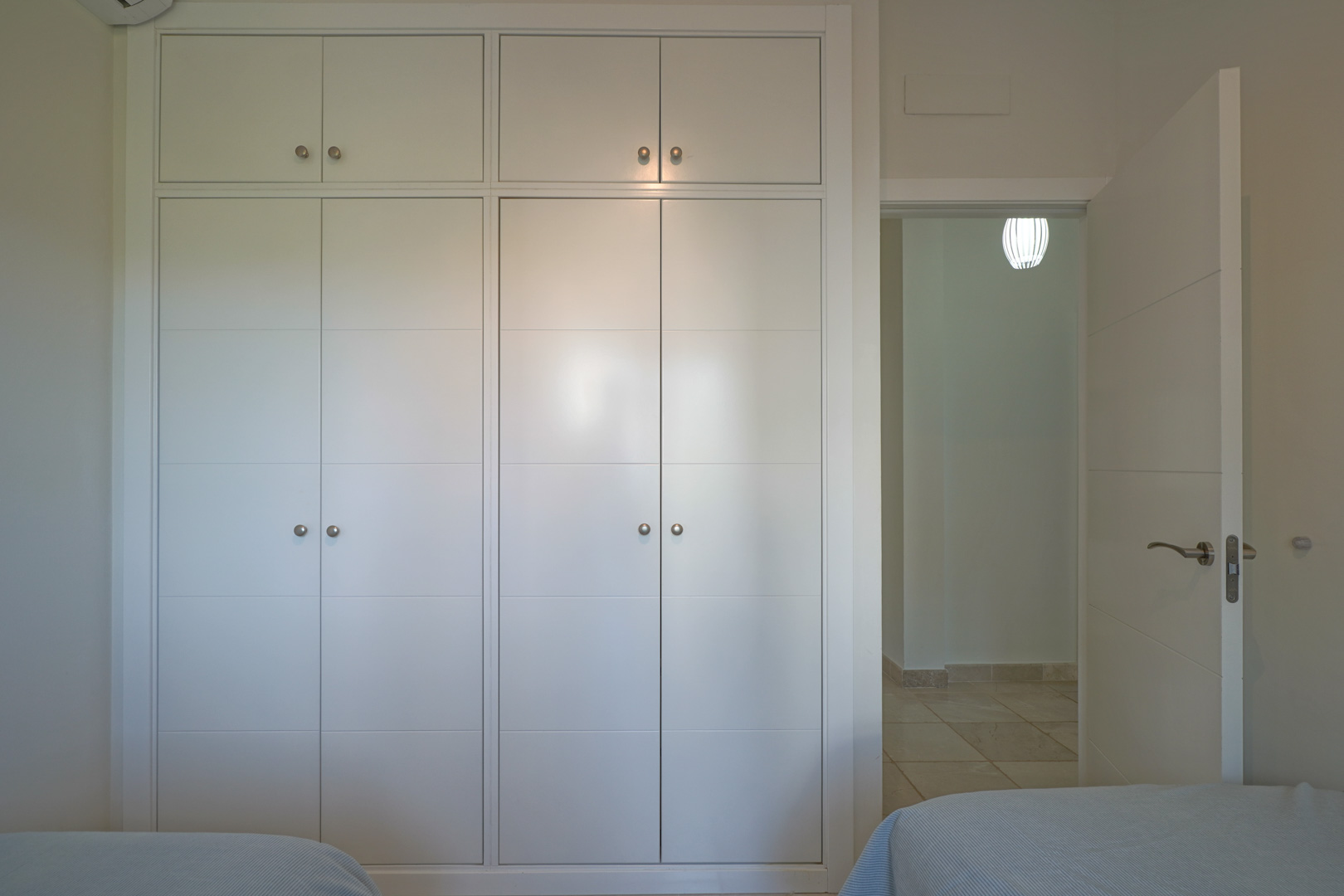 Floor-to-ceiling white built-in wardrobe with silver knobs and upper storage, located next to the bedroom entrance with views of the tiled hallway.