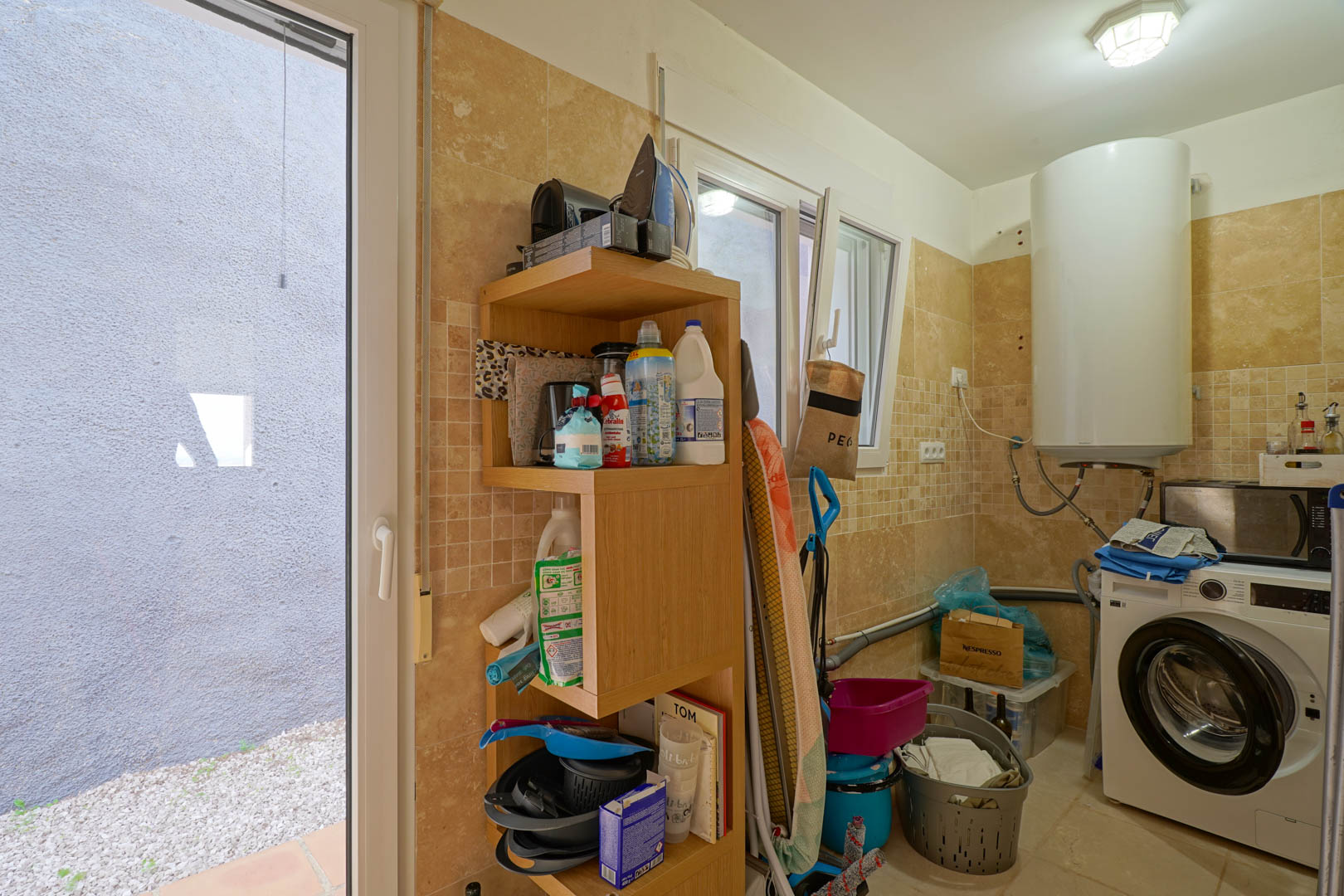 Functional laundry room featuring cream-toned tiled walls, an electric water heater, washing machine, wooden storage unit, and direct access to the outdoor area.