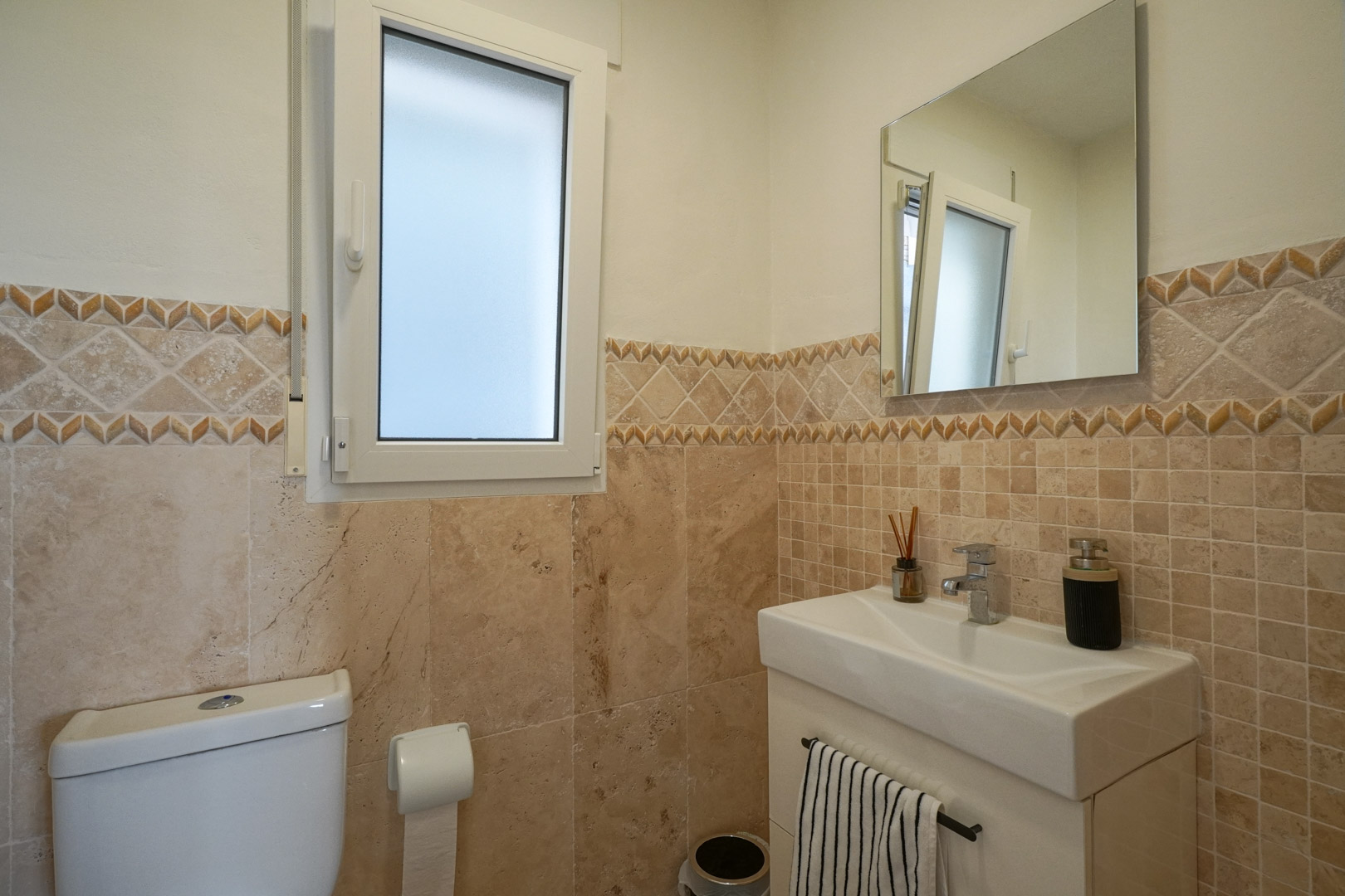 Guest toilet featuring earth-toned tiling with a decorative border, a modern white vanity, and a frosted glass window for privacy and light.