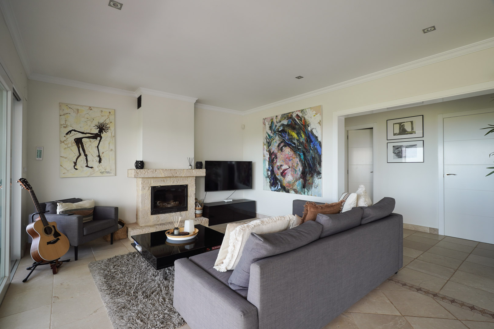 Living area featuring a natural stone fireplace, grey seating, and tiled floors with decorative borders. Contemporary art and recessed lighting define this elegant space.