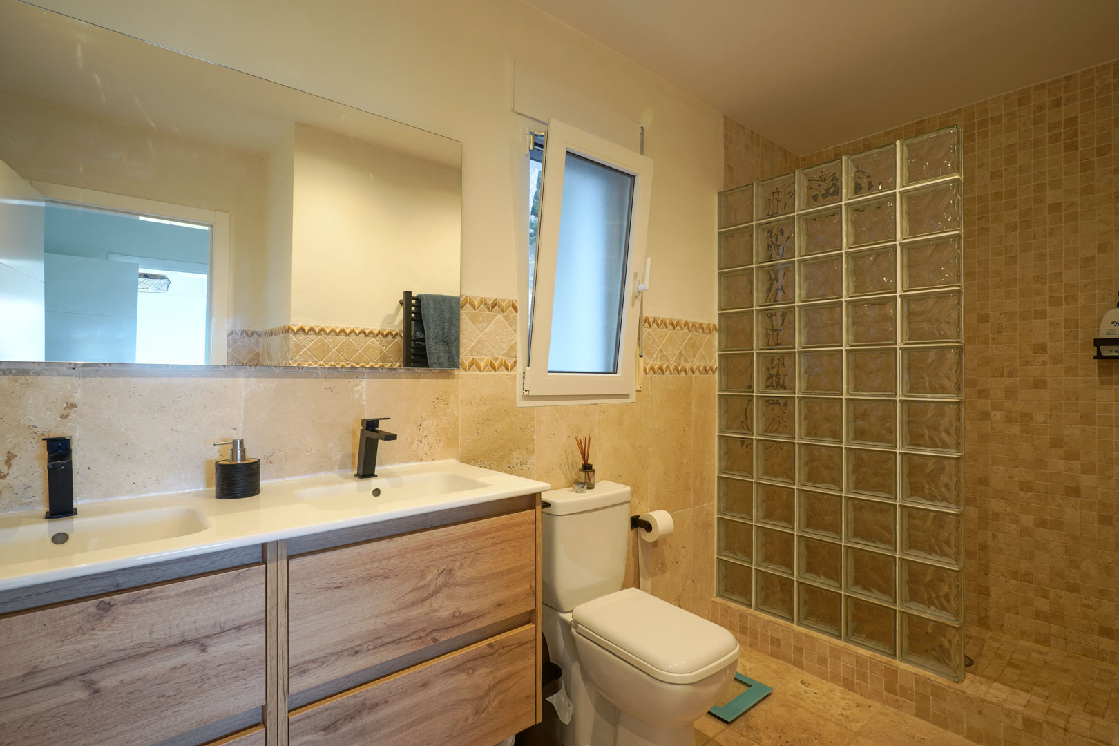 Modern bathroom featuring a wooden double vanity with black faucets and a walk-in shower with a glass block partition.