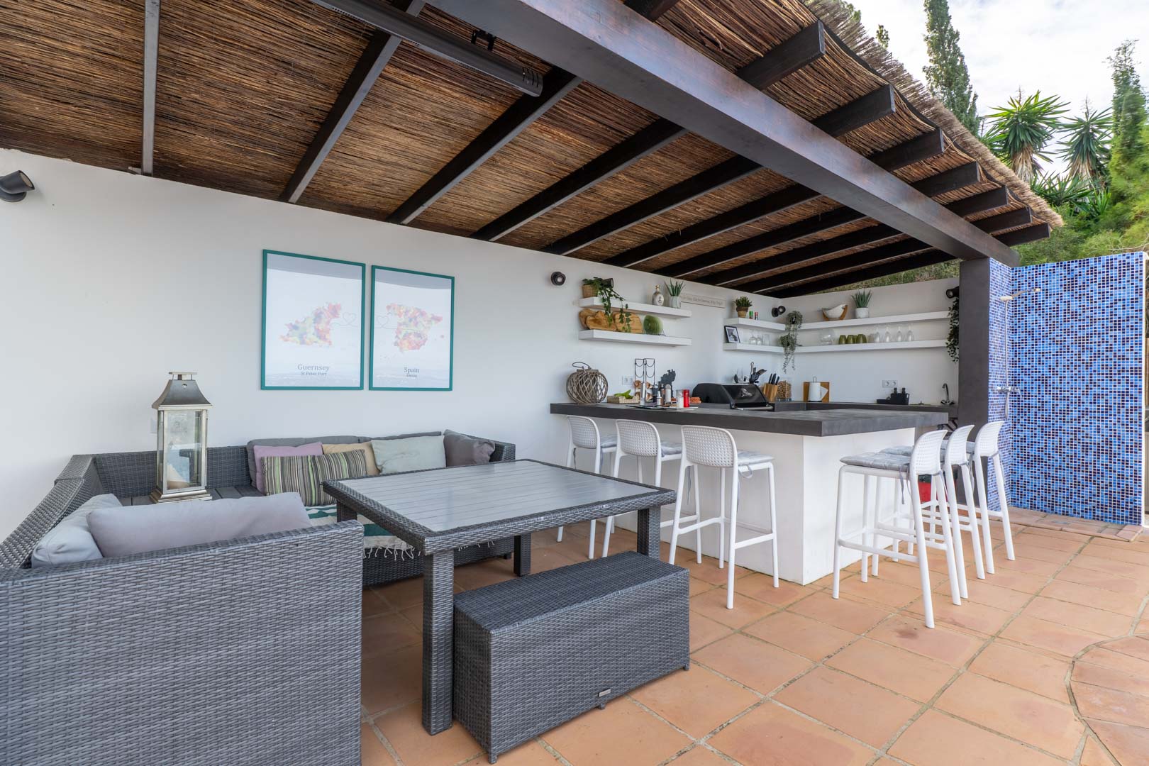 Outdoor kitchen and lounge area under a wooden pergola with reed roofing, featuring a breakfast bar with white stools, grey rattan furniture, and terracotta flooring.
