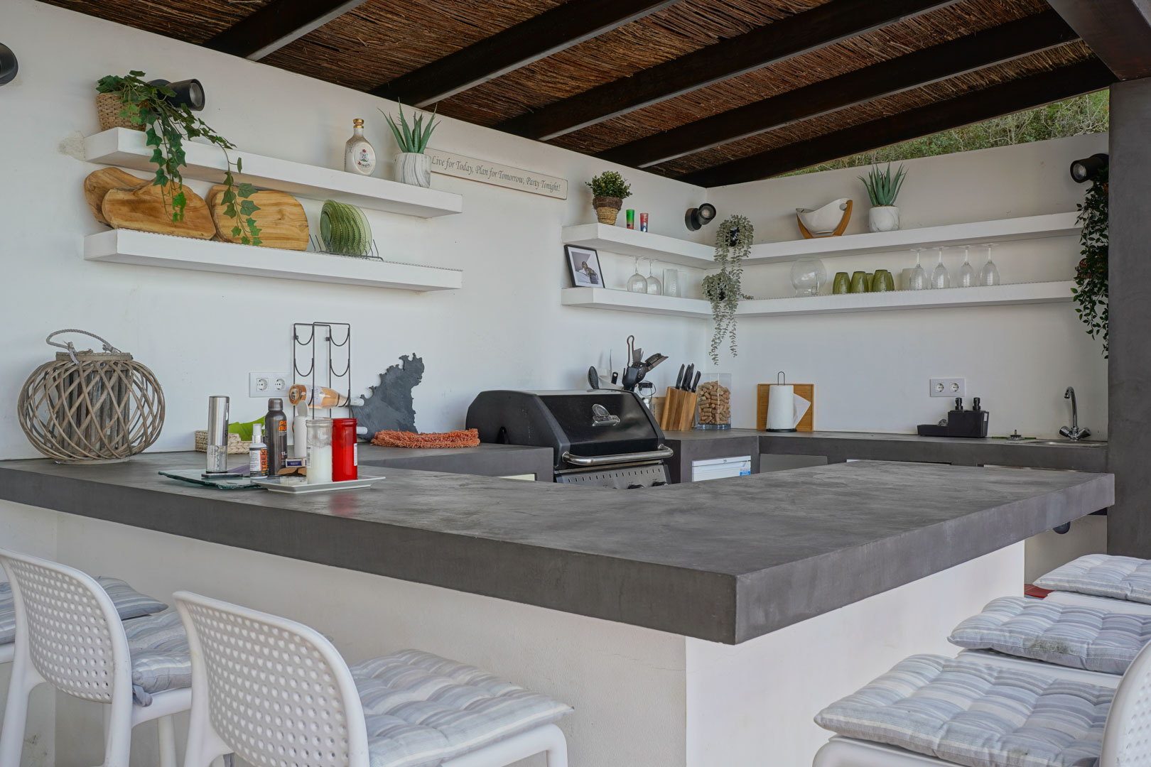 Outdoor summer kitchen featuring grey concrete countertops, an integrated gas grill, open shelving, and a rustic thatched ceiling with wooden beams.