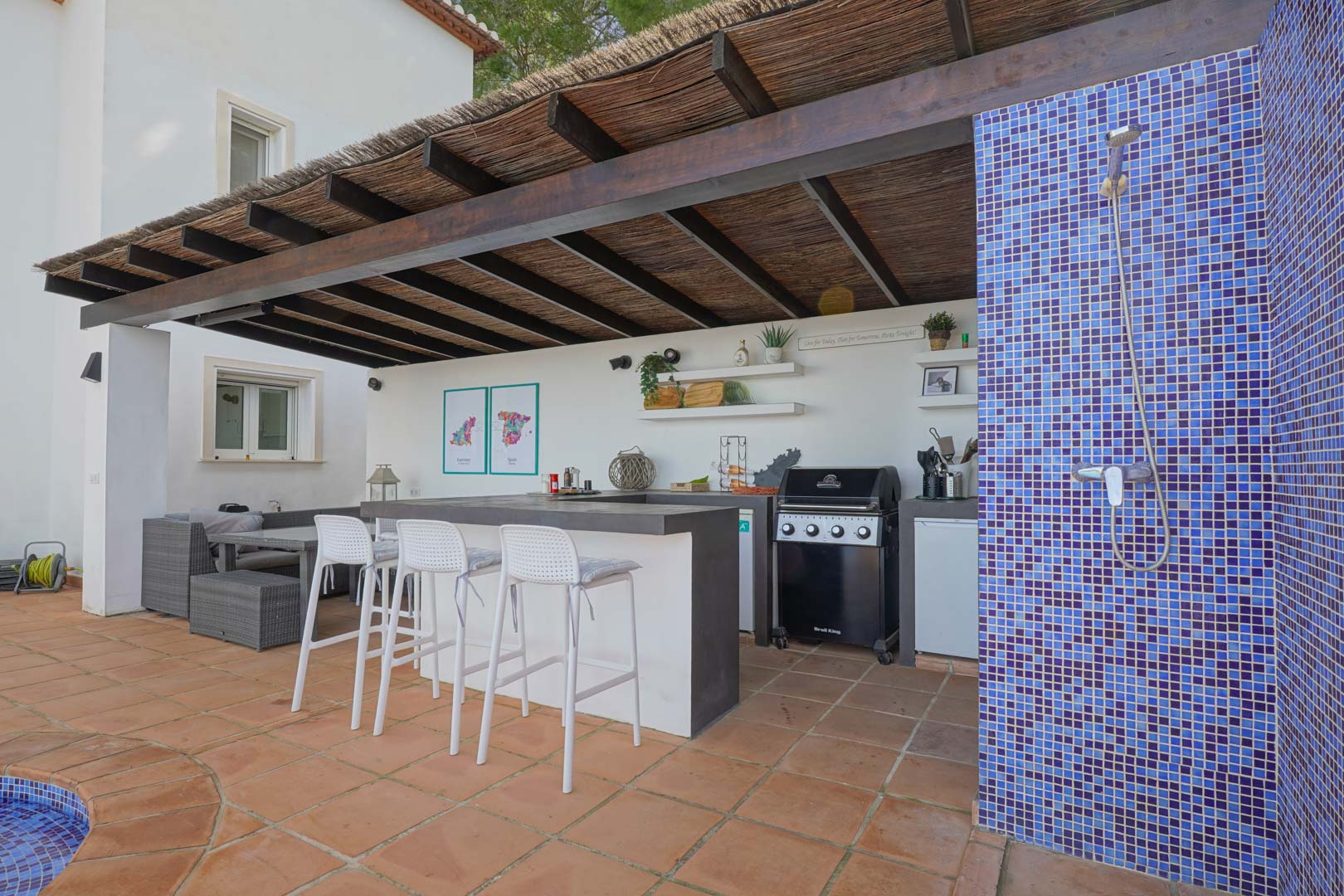 Outdoor summer kitchen under a wooden pergola with a heather roof, featuring a gas grill, bar seating, and an adjacent blue mosaic tile shower.