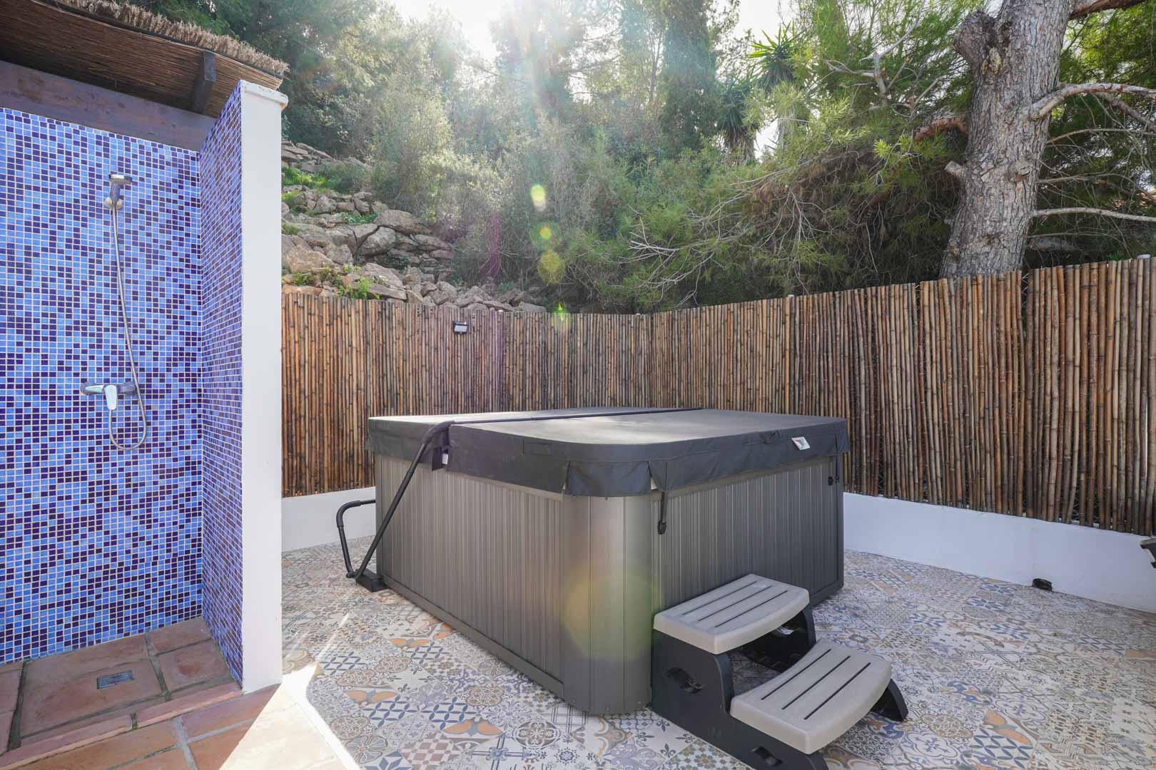 Outdoor wellness area featuring a covered jacuzzi, blue mosaic tile shower, and patterned hydraulic flooring, enclosed by natural bamboo fencing.