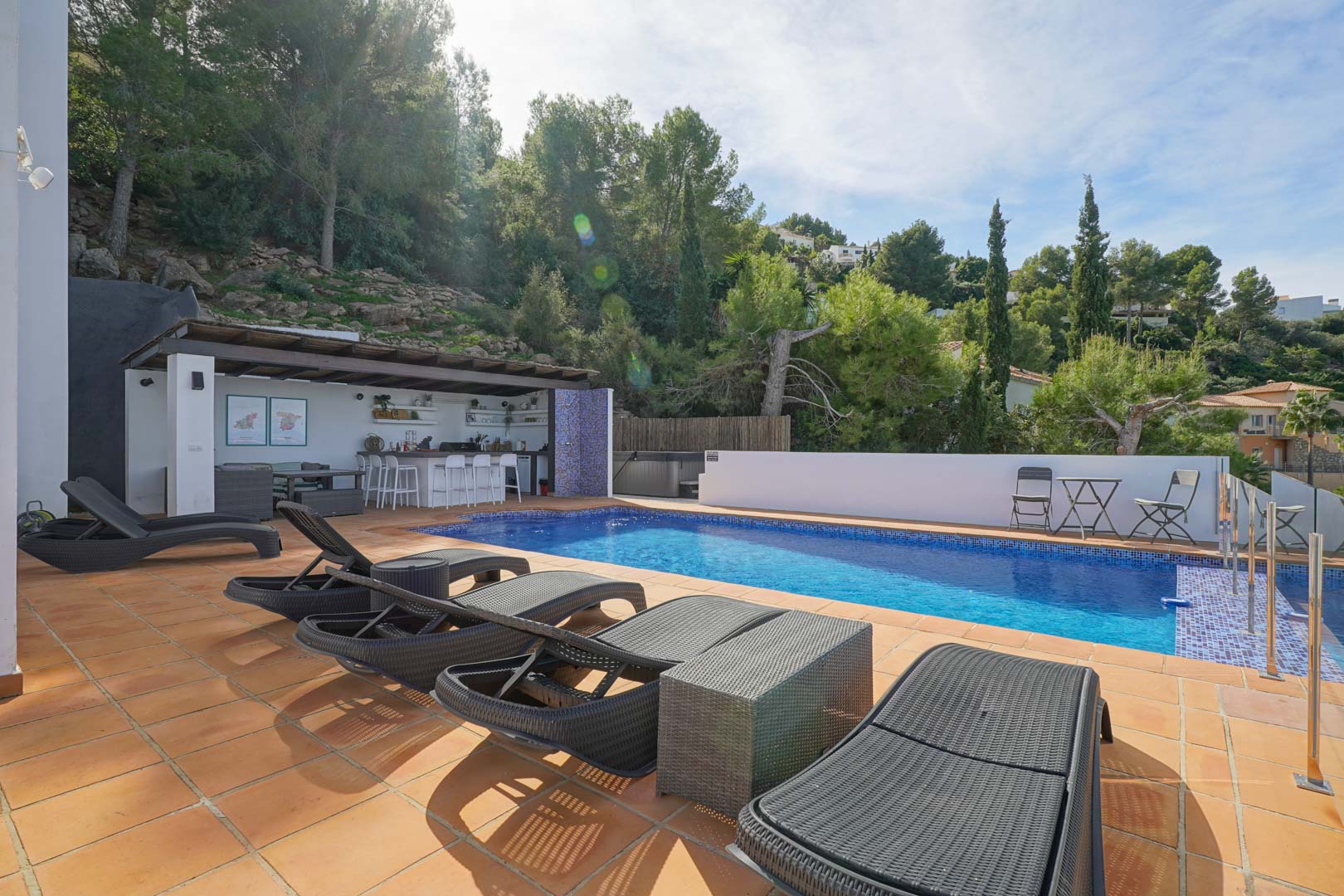 Private pool area with terracotta tiling, dark rattan sun loungers, and a shaded summer bar with seating, set against a lush Mediterranean pine hillside.