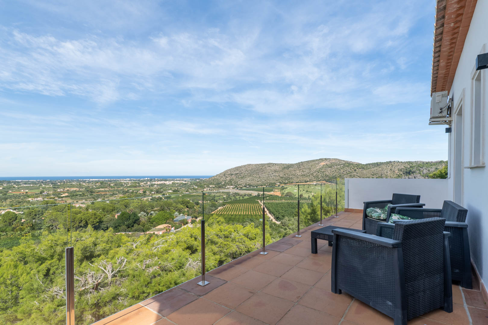 Upper terrace featuring a glass balustrade, dark wicker furniture, and panoramic views of the Mediterranean Sea, the golf course, and the surrounding coastal landscape.