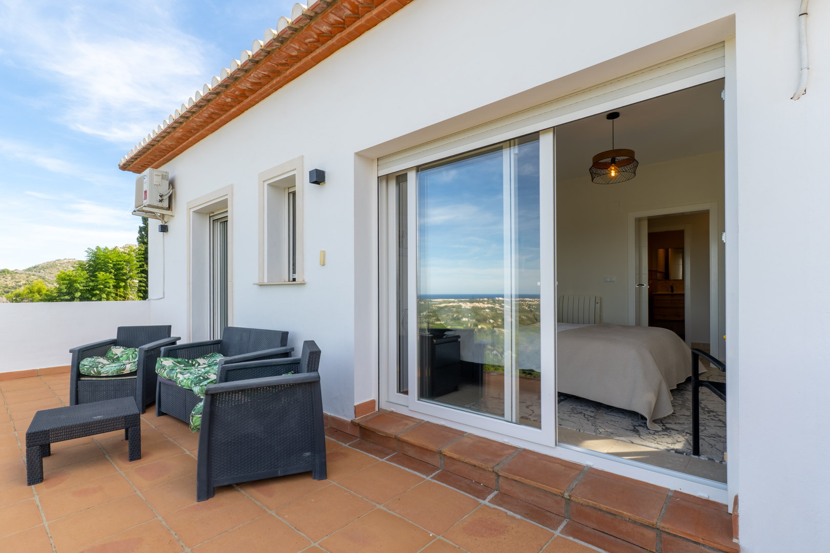 Upper terrace featuring terracotta tiling and outdoor seating, with large sliding glass doors providing direct bedroom access and panoramic coastal views.