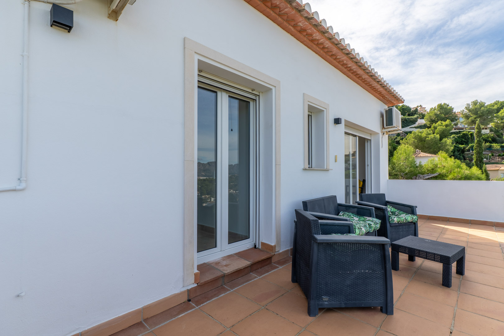 Upper terrace featuring terracotta tiling, dark wicker outdoor seating, and direct access to the bedrooms through white-framed glass doors.