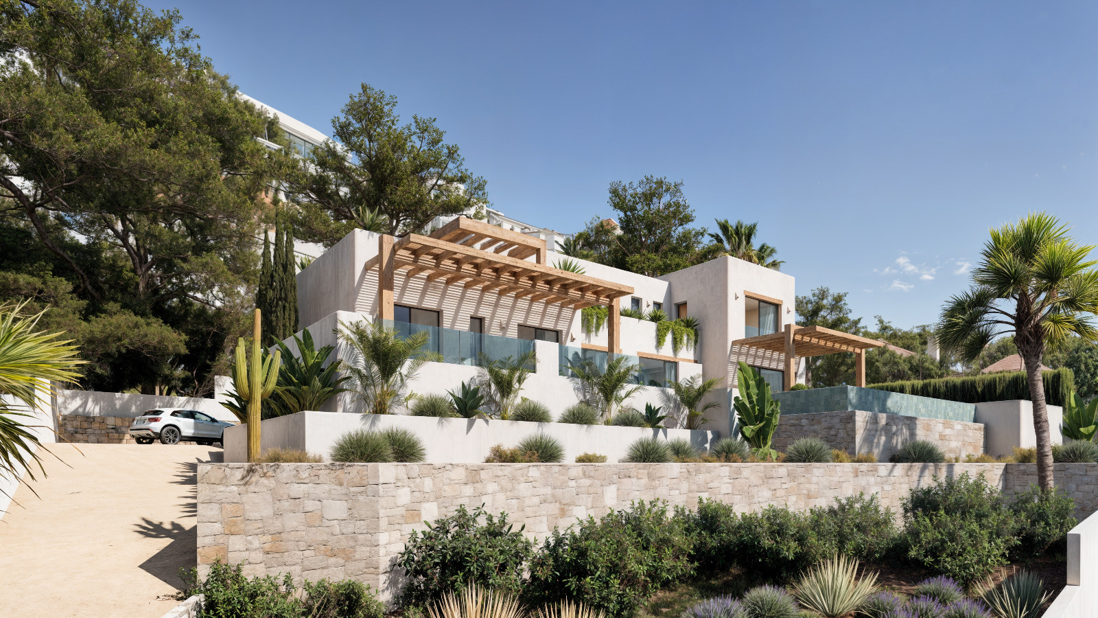 Exterior view of the Ibicenco-style villa featuring white facades, wooden pergolas, and natural stone walls integrated into a Mediterranean garden with palms.