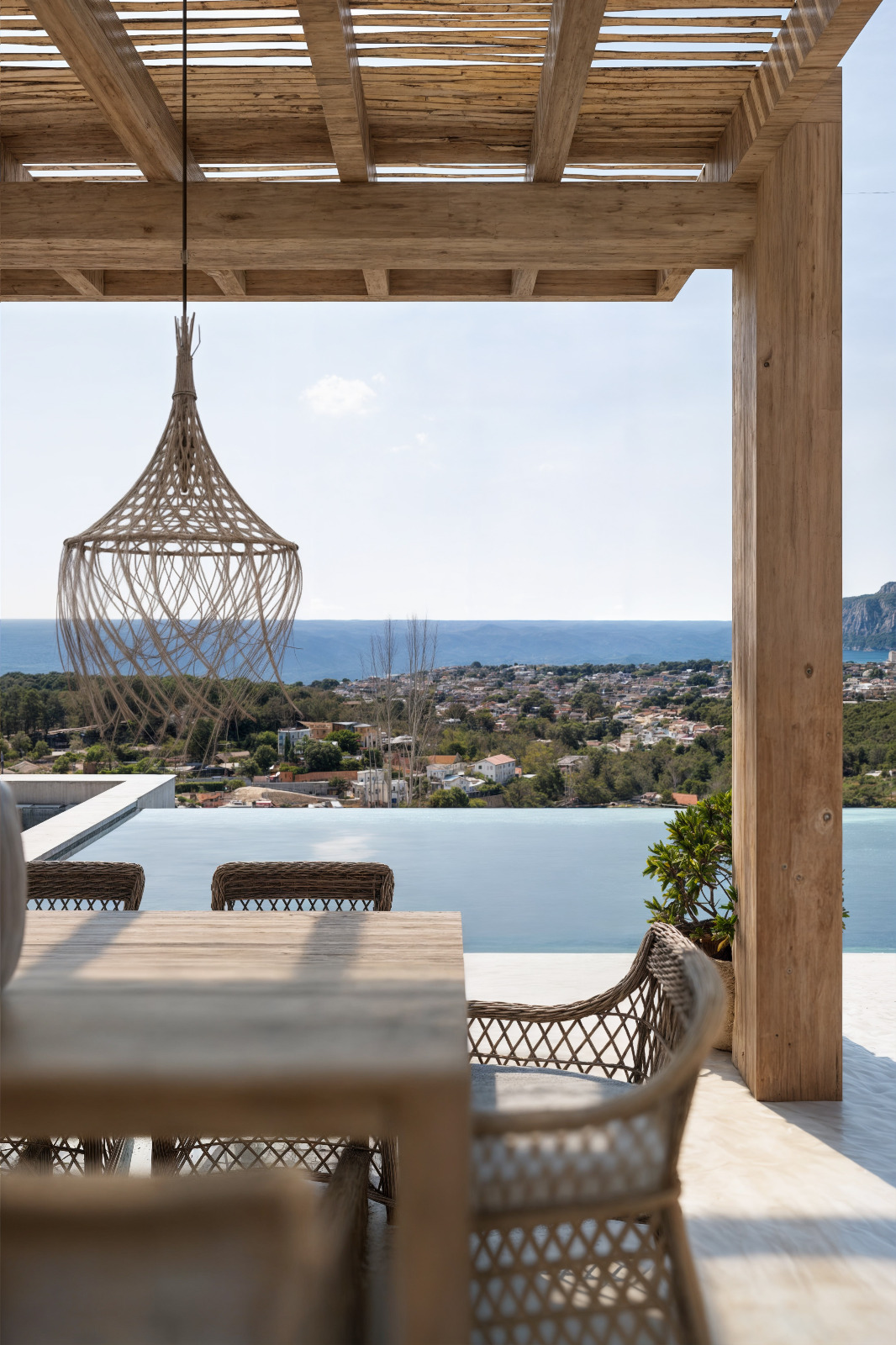 Outdoor terrace featuring a wooden pergola, wicker dining furniture, and a woven pendant light overlooking the infinity pool and Mediterranean Sea.