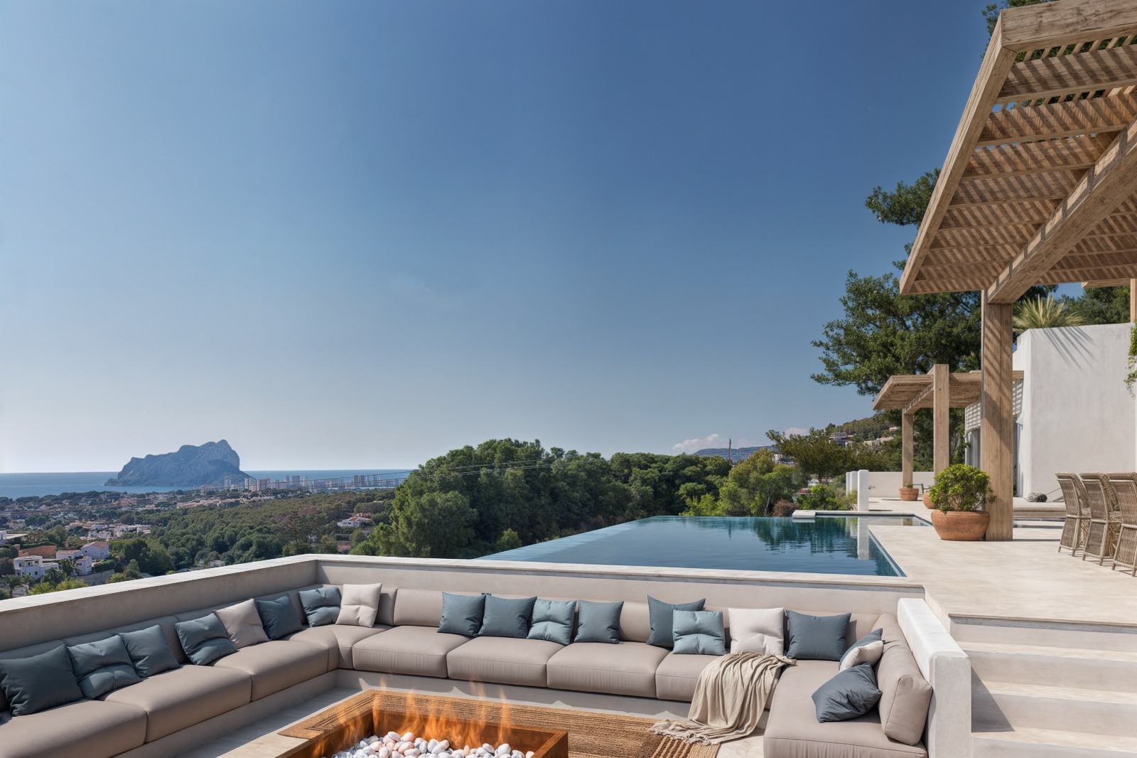 Sunken lounge area featuring a central fire pit and perimeter seating, overlooking an infinity pool and the Mediterranean coastline with the Peñón de Ifach.
