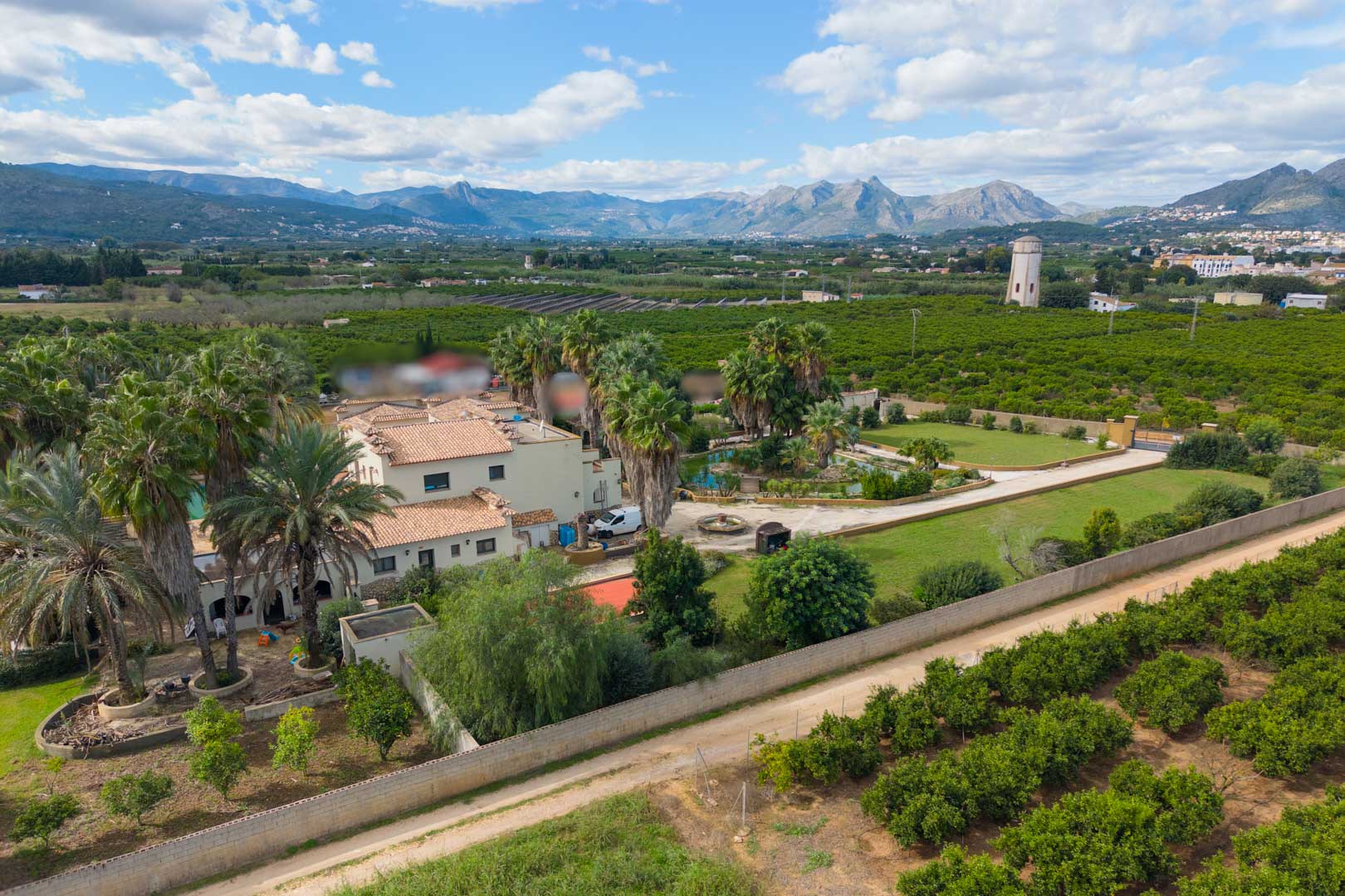 Aerial perspective of the estate featuring the main villa, palm-filled gardens, and a pond, bordered by orange groves with distant mountain views.