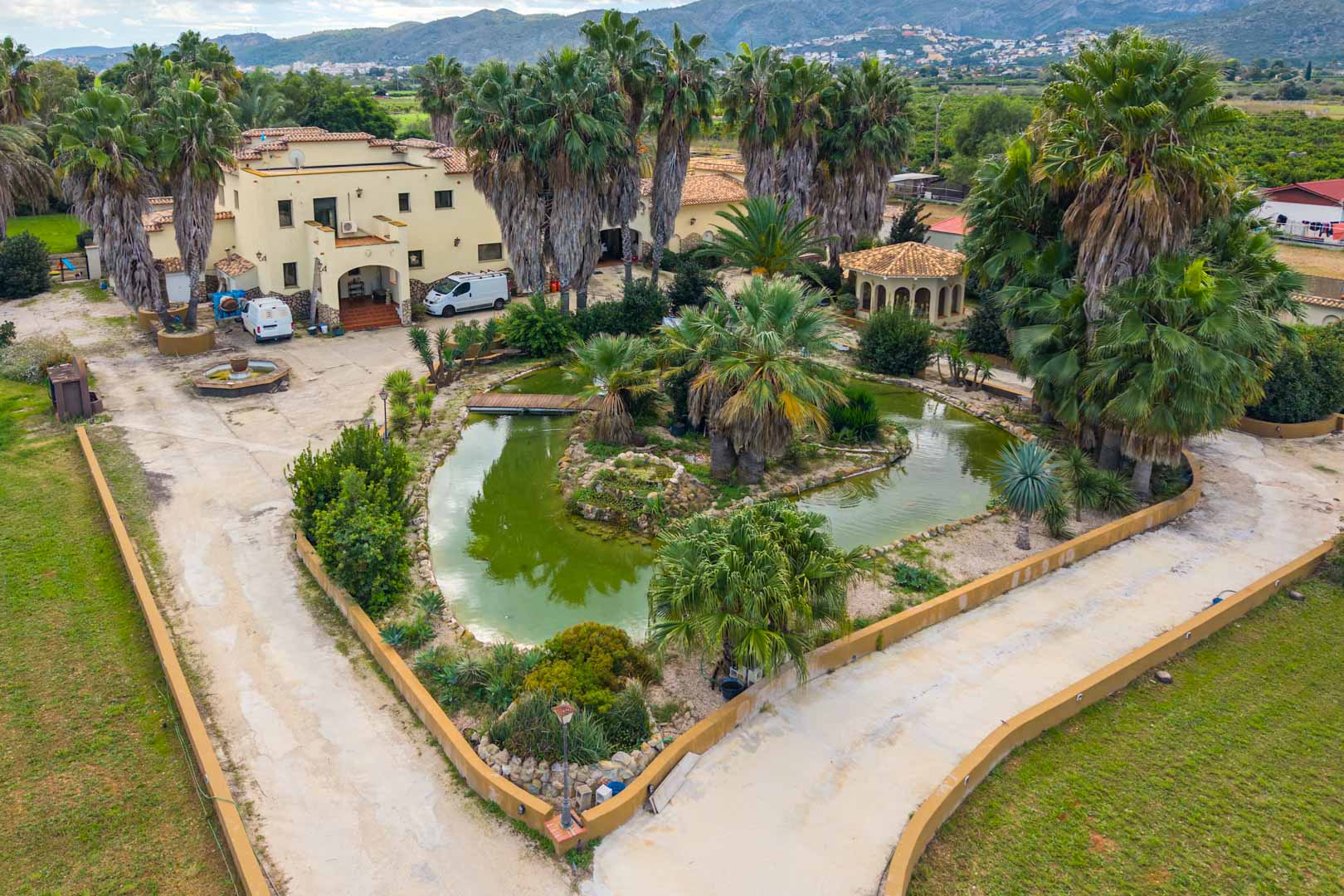 Aerial view of the main entrance featuring a large ornamental pond with a wooden bridge, surrounded by mature palm trees and the Mediterranean villa.