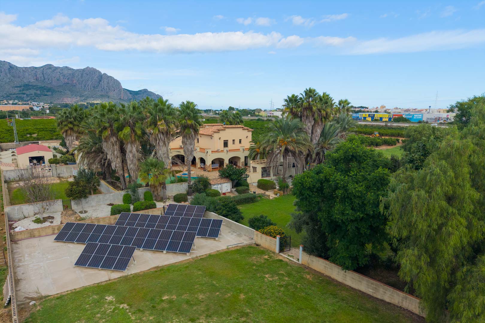 Aerial view showing the extensive solar panel installation, manicured lawns, and the Mediterranean villa framed by mature palm trees and mountains.