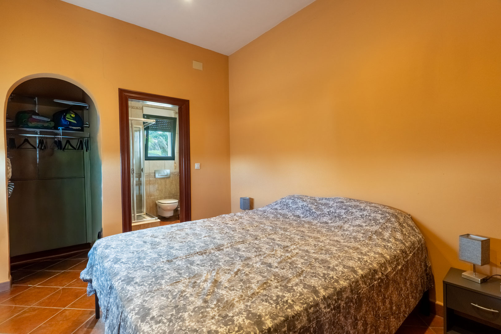 Bedroom with terracotta flooring and warm ochre walls, featuring an arched walk-in closet and an en-suite bathroom with a shower.
