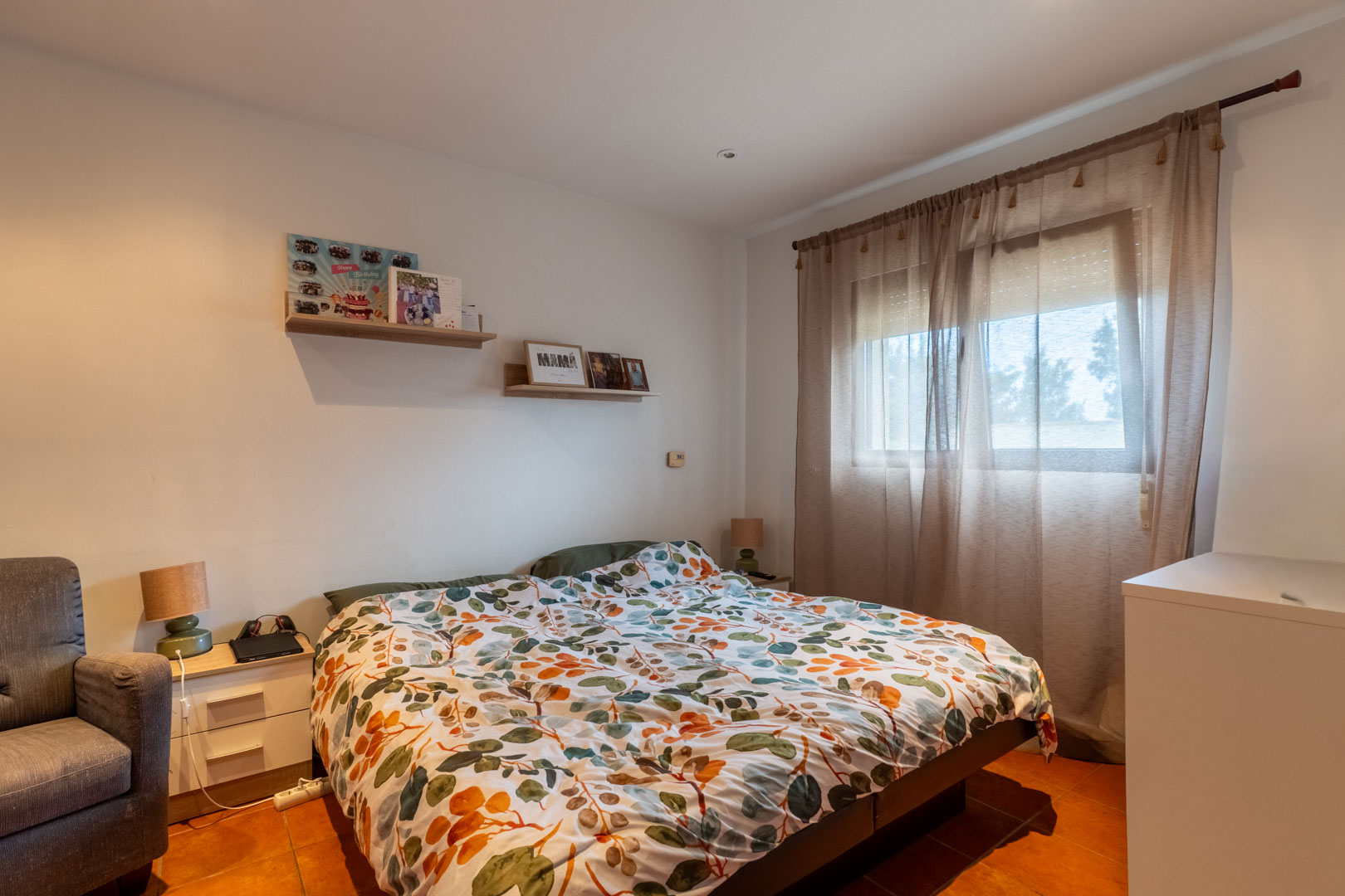 Bright bedroom featuring terracotta tile flooring, white walls, and wooden floating shelves. A window with translucent curtains and a roller shutter provides natural light.
