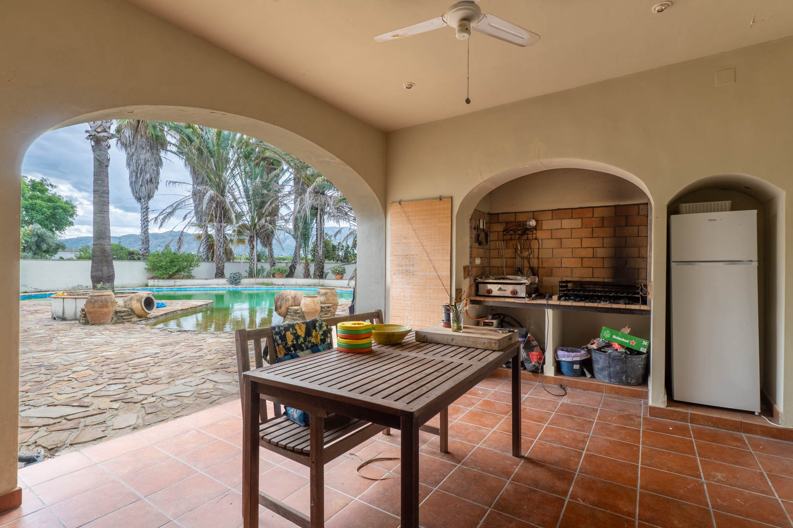 Covered porch featuring a built-in brick barbecue, wooden dining set, and a large archway overlooking the swimming pool and palm-lined gardens.