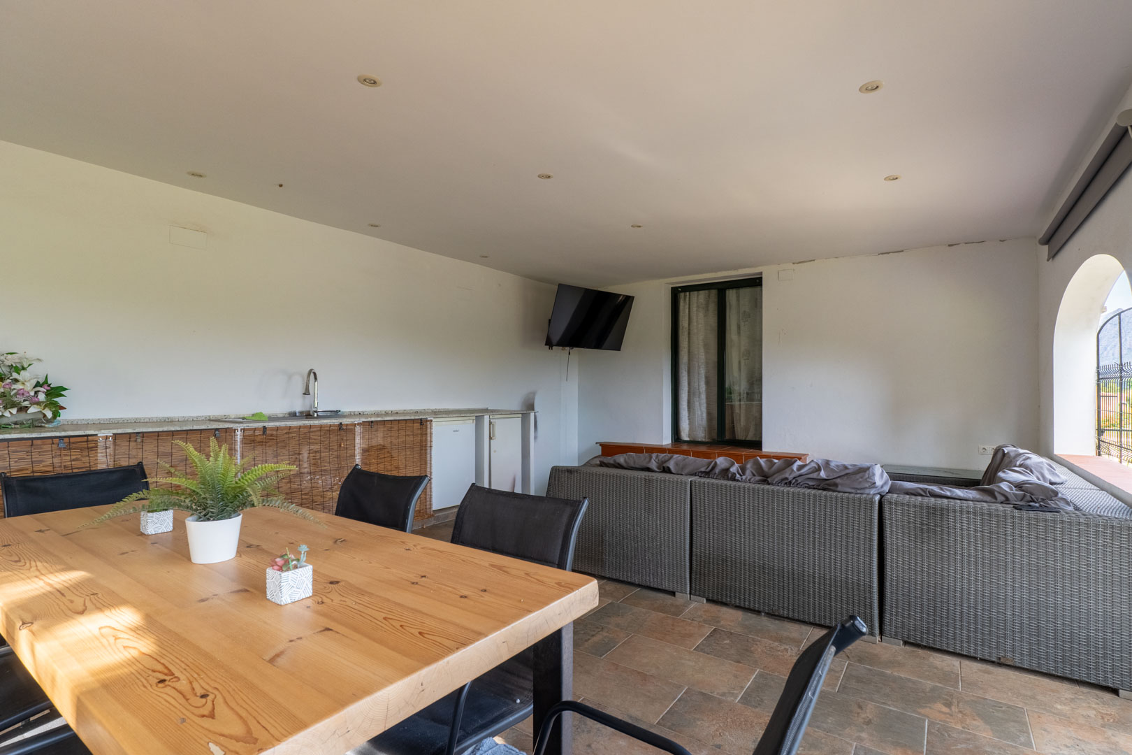 Covered terrace featuring a grey wicker sectional sofa, a solid wood dining table, and an outdoor kitchenette. The space includes recessed ceiling lights and tiled flooring.