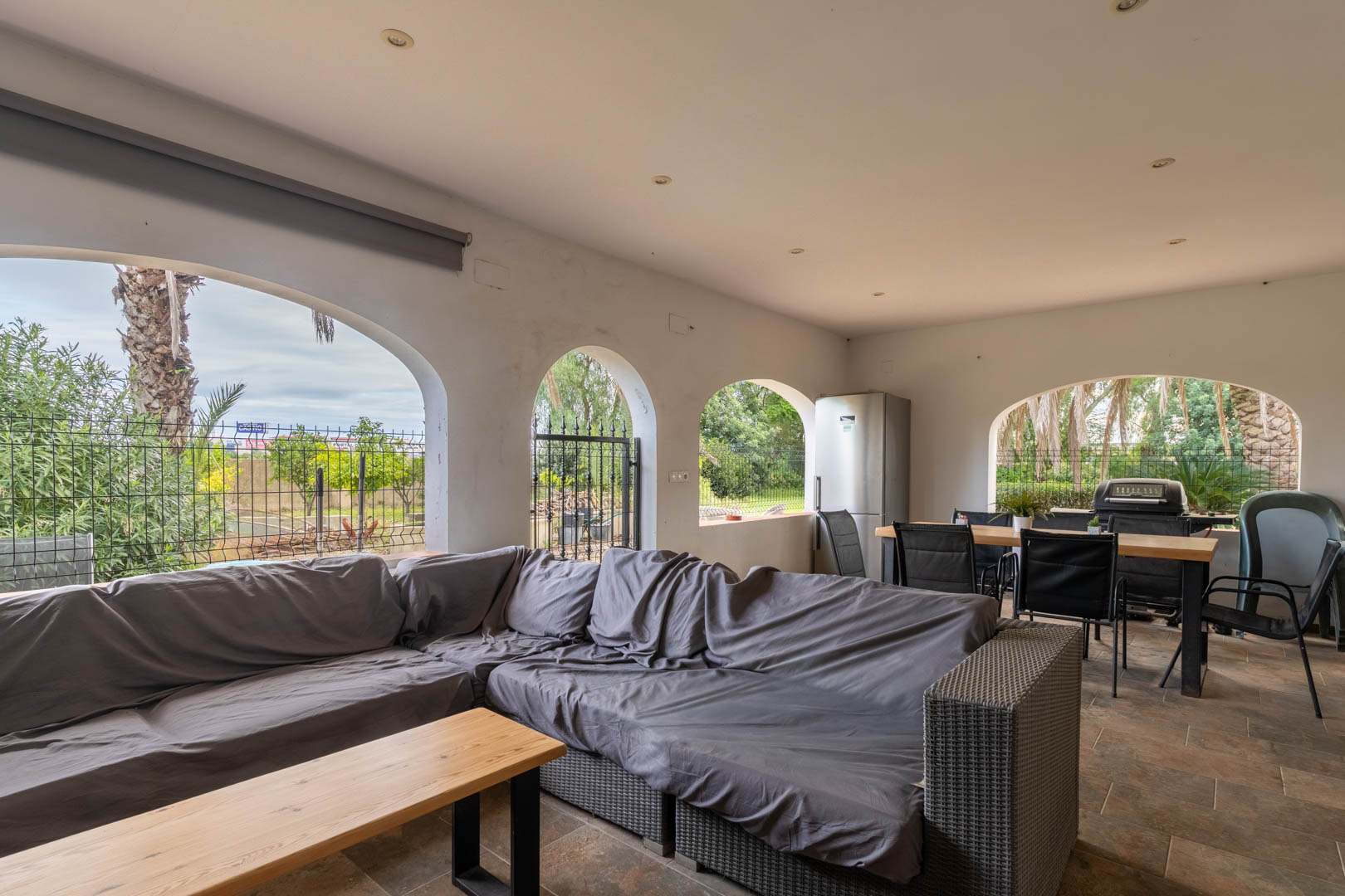Covered terrace featuring Mediterranean arches, a grey lounge area, and an outdoor dining space with a barbecue and fridge. Tiled flooring and recessed lighting.